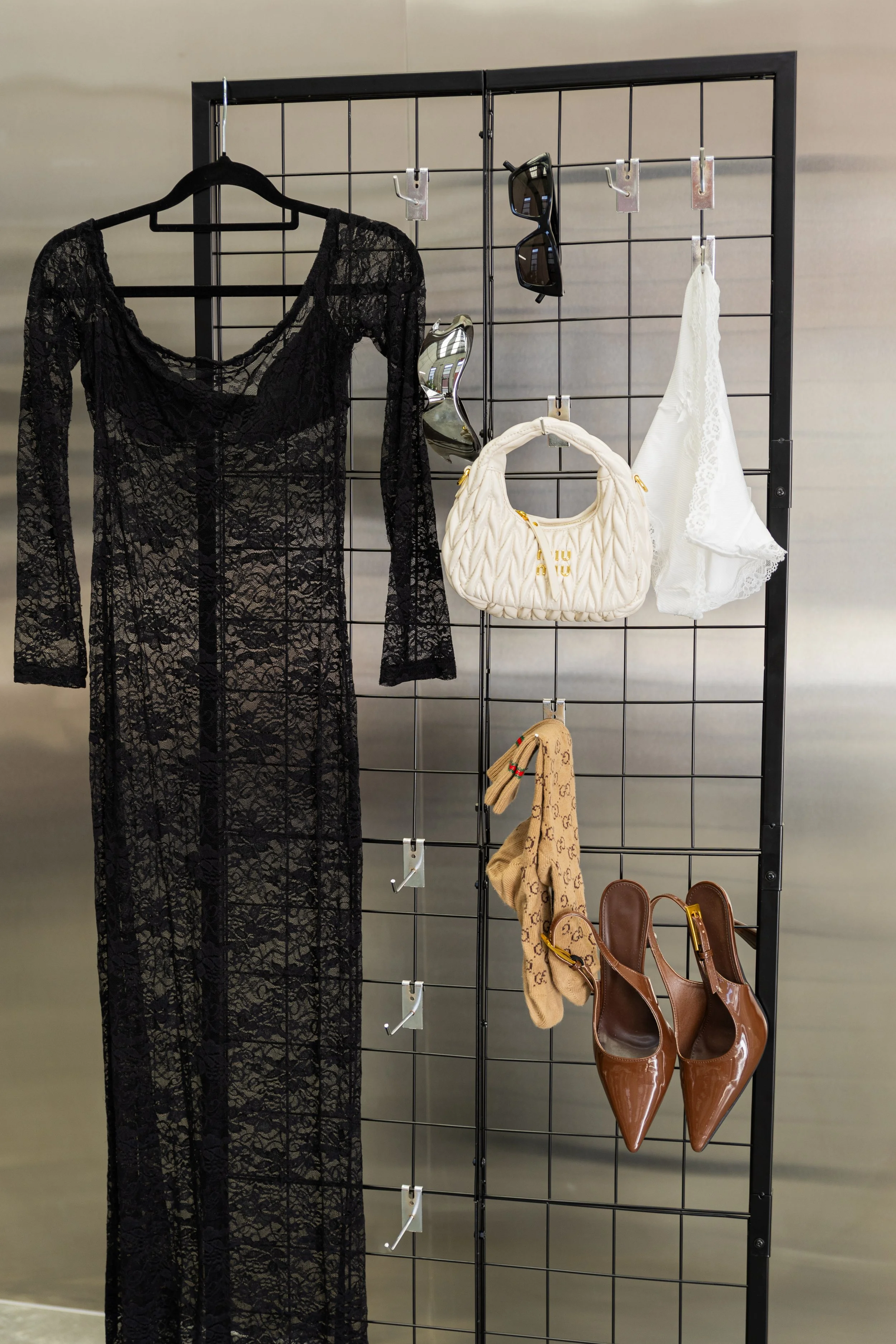 Clothing and accessories display rack with black lace dress, sunglasses, white purse, beige scarf, white lace garment, and brown high heel shoes.