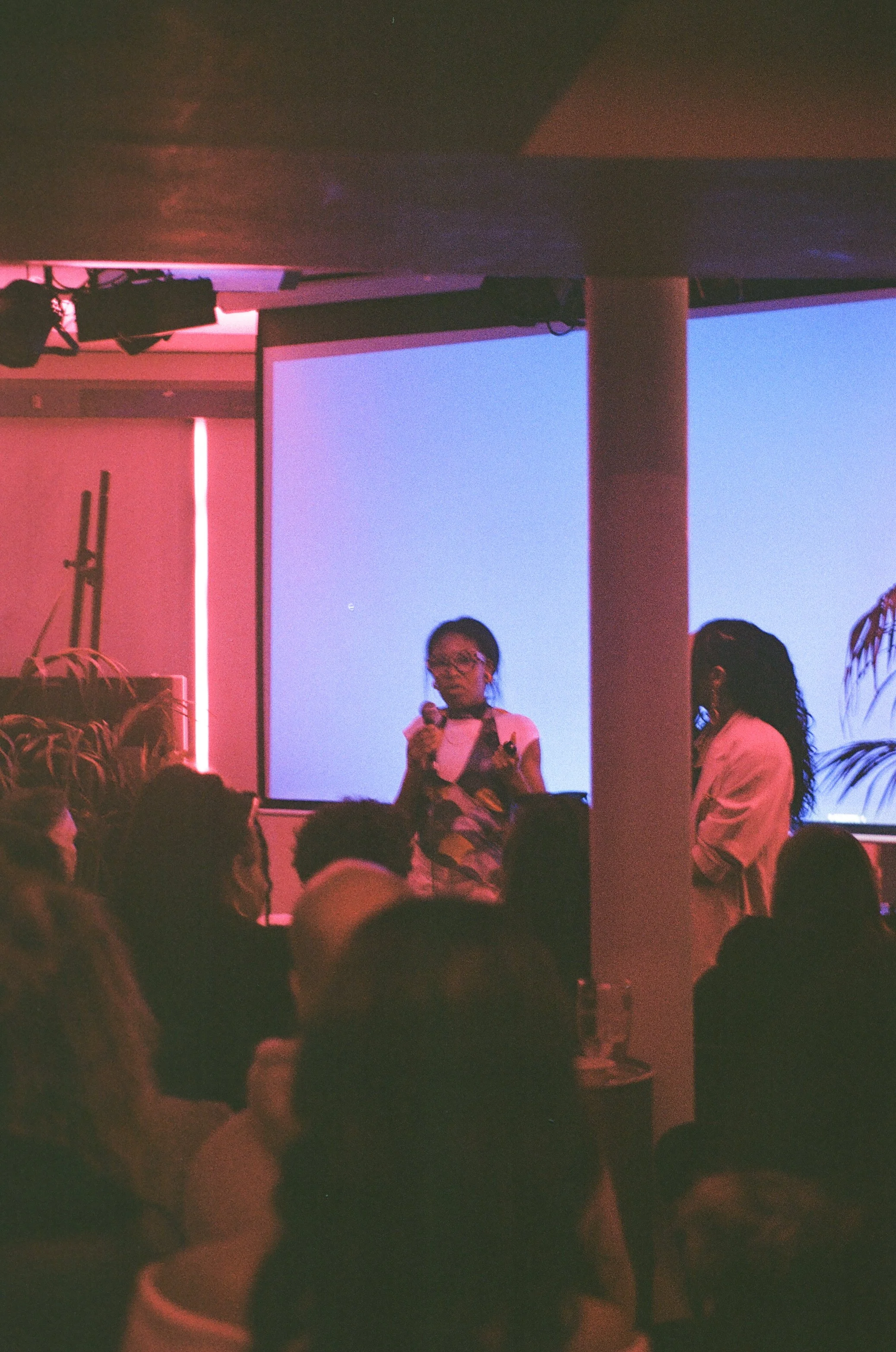 Victory, editor of Bia!, is speaking into a microphone on a stage with a large blue screen behind her, while Rema, co-editor of Issue 2, stands nearby. The audience is seated in a dimly lit room with pink neon accent lighting.