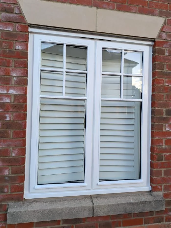 A white-framed sliding glass window with partially closed white blinds, set in a red brick wall with a concrete sill.