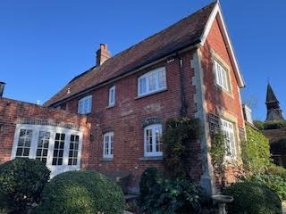 A large brick house with a sloped roof and multiple windows, surrounded by greenery.