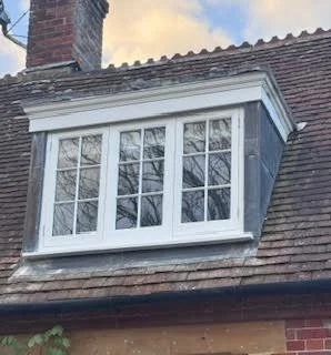 The photograph shows a traditional brick house roof with a large white bay window on the upper floor, featuring multiple panes and a slightly sloped roof.