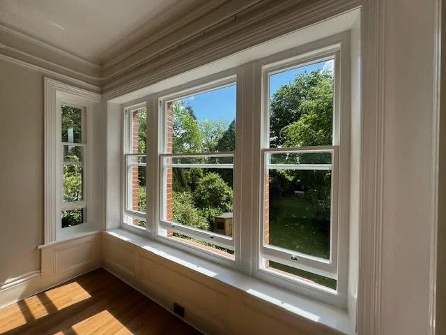 Three large windows overlooking a lush green garden with trees, allowing natural light into a room with wooden flooring.