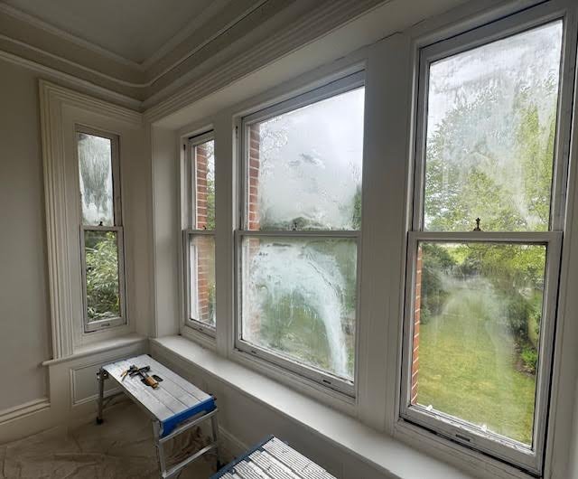 Interior view of a room with large windows and condensation, overlooking greenery. A workbench with tools is set up by the windows, suggesting maintenance or cleaning work in progress.