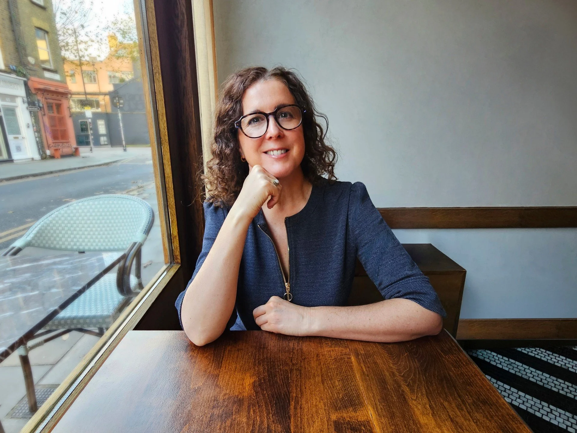 A woman with curly brown hair, glasses, wearing a dark blue zip-up top with a gold zipper, sitting at a wooden table near a large window inside a cafe. She is smiling and resting her chin on her left hand.