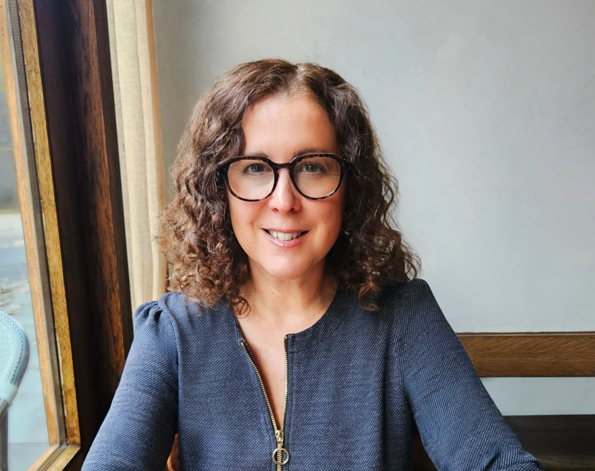 A woman with curly brown hair, wearing glasses and a dark blue zippered top, smiling for a selfie indoors near a window.