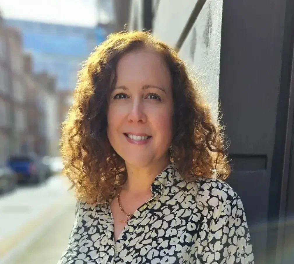 A woman with curly brown hair smiling outdoors in sunlight, wearing a patterned blouse with a black and white floral design.