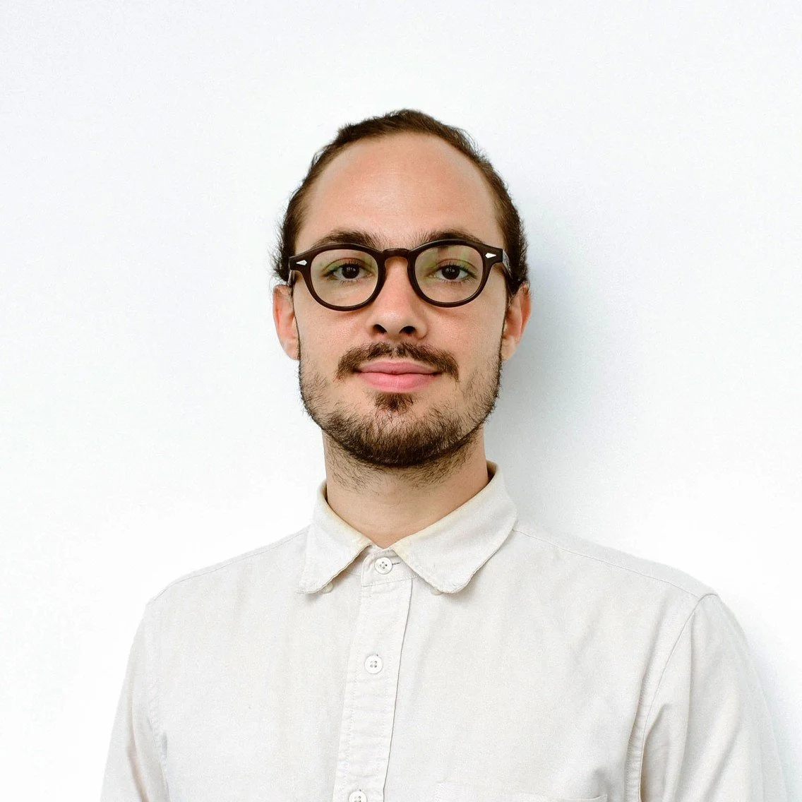 A young man with glasses, a beard, and long hair tied back, wearing a white button-up shirt, standing in front of a plain white wall.