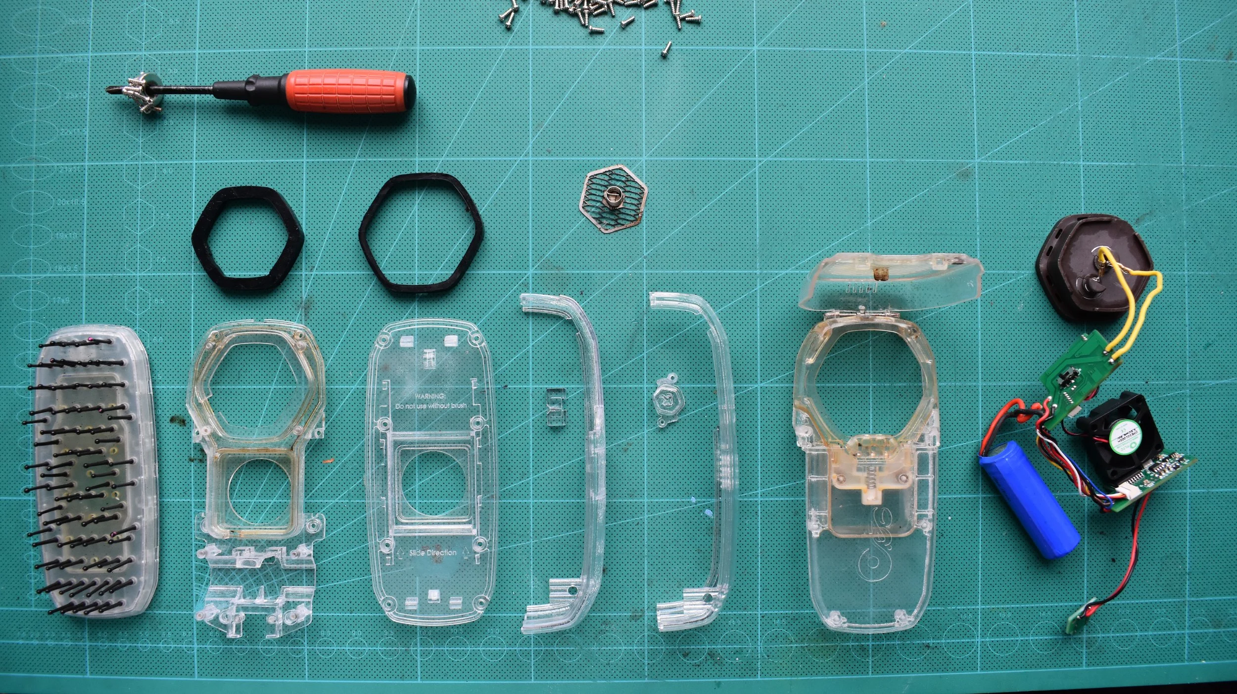 Disassembled electronic device parts laid out on a green cutting mat, including plastic shells, screws, wires, a small fan, and a screwdriver.
