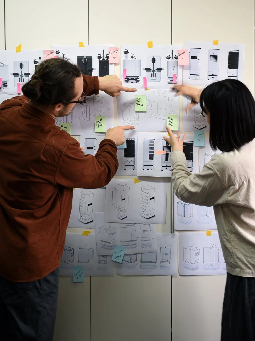 Two people collaborating on a design board with sketches, photos, and sticky notes for a technology product, planning features and layout.