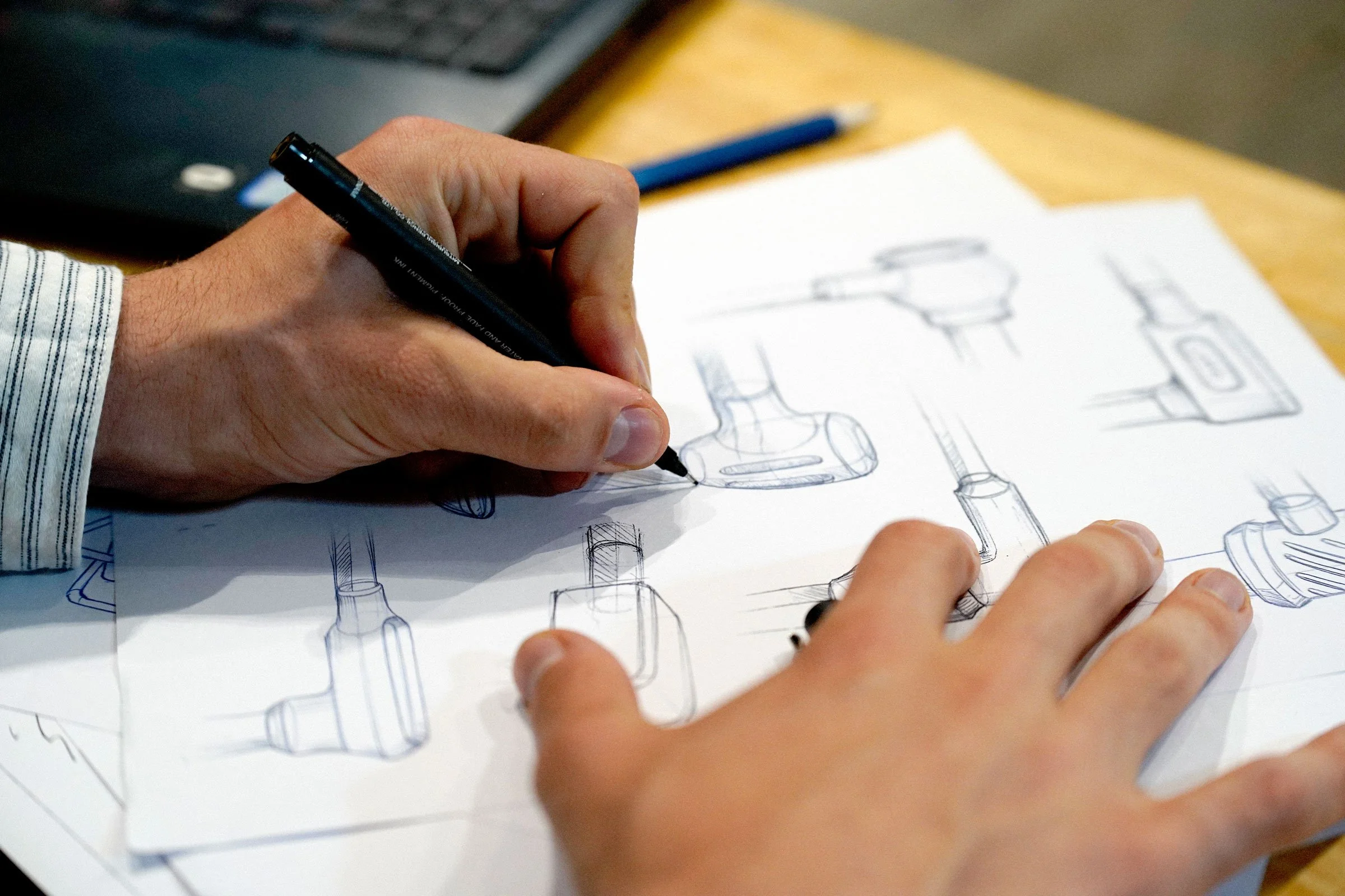 Person sketching mechanical parts on paper with a black pen alongside a blue pen, with a laptop in the background.