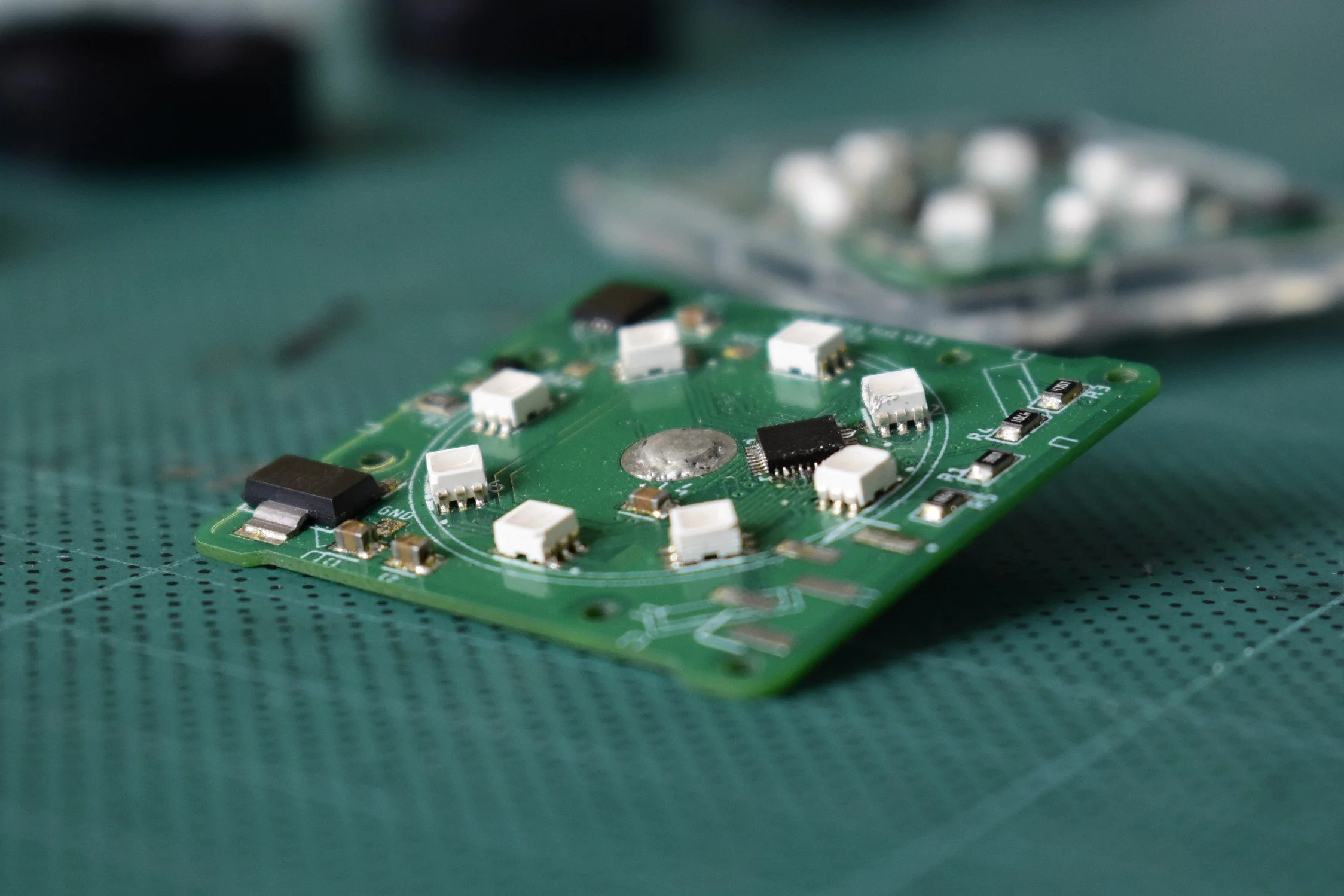 A close-up of a green circuit board with various electronic components, resting on a textured green surface.