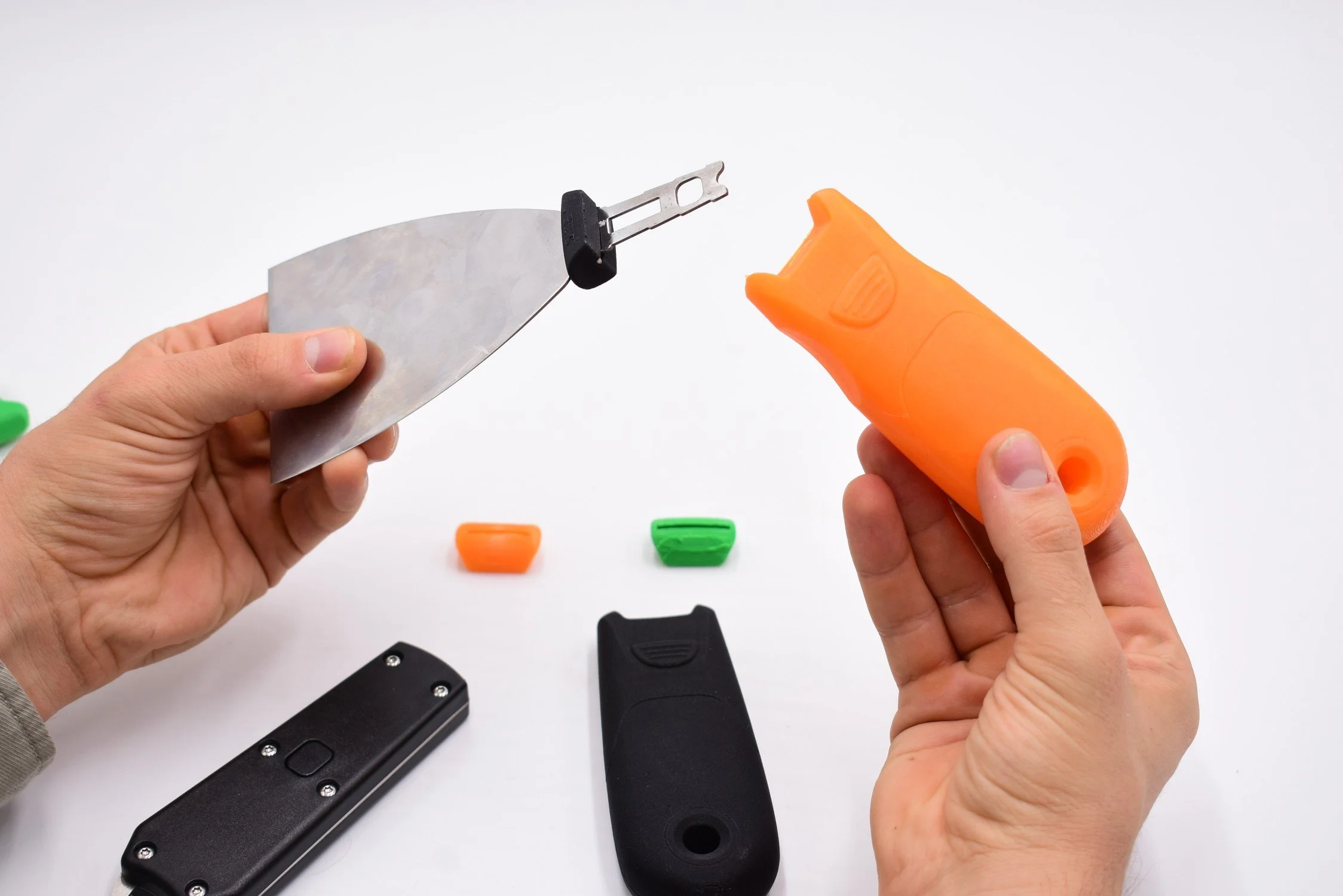 Hands holding a small razor blade with an orange plastic handle, with colorful small objects and black devices on a white background.
