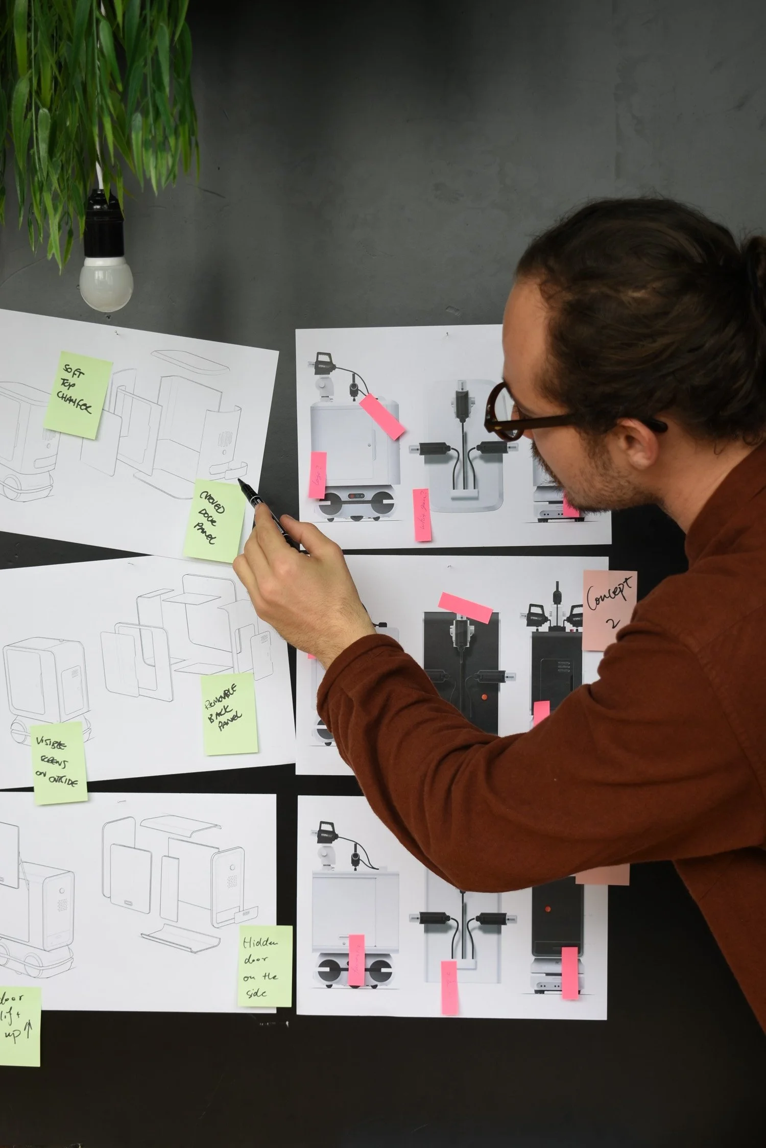 A man with glasses and brown hair, wearing a brown jacket, is looking at design sketches and diagrams posted on a black table surface. The sketches include wireframe diagrams and concept images with sticky notes attached. There is a potted plant with green leaves and a white light bulb hanging from a black socket in the top left corner.