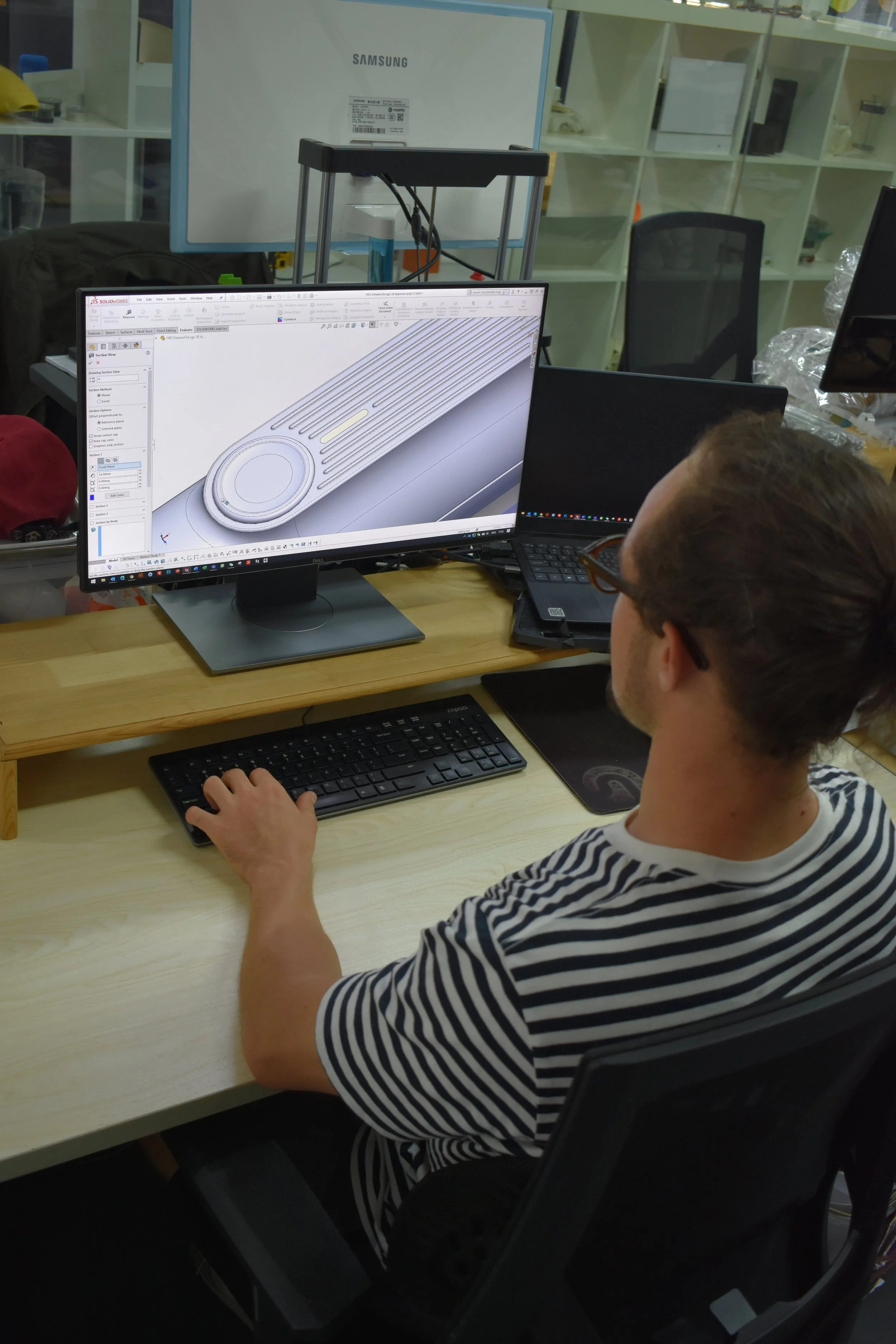 A person working at a computer with design software displaying a 3D model of a mechanical part, in a workshop or office environment.