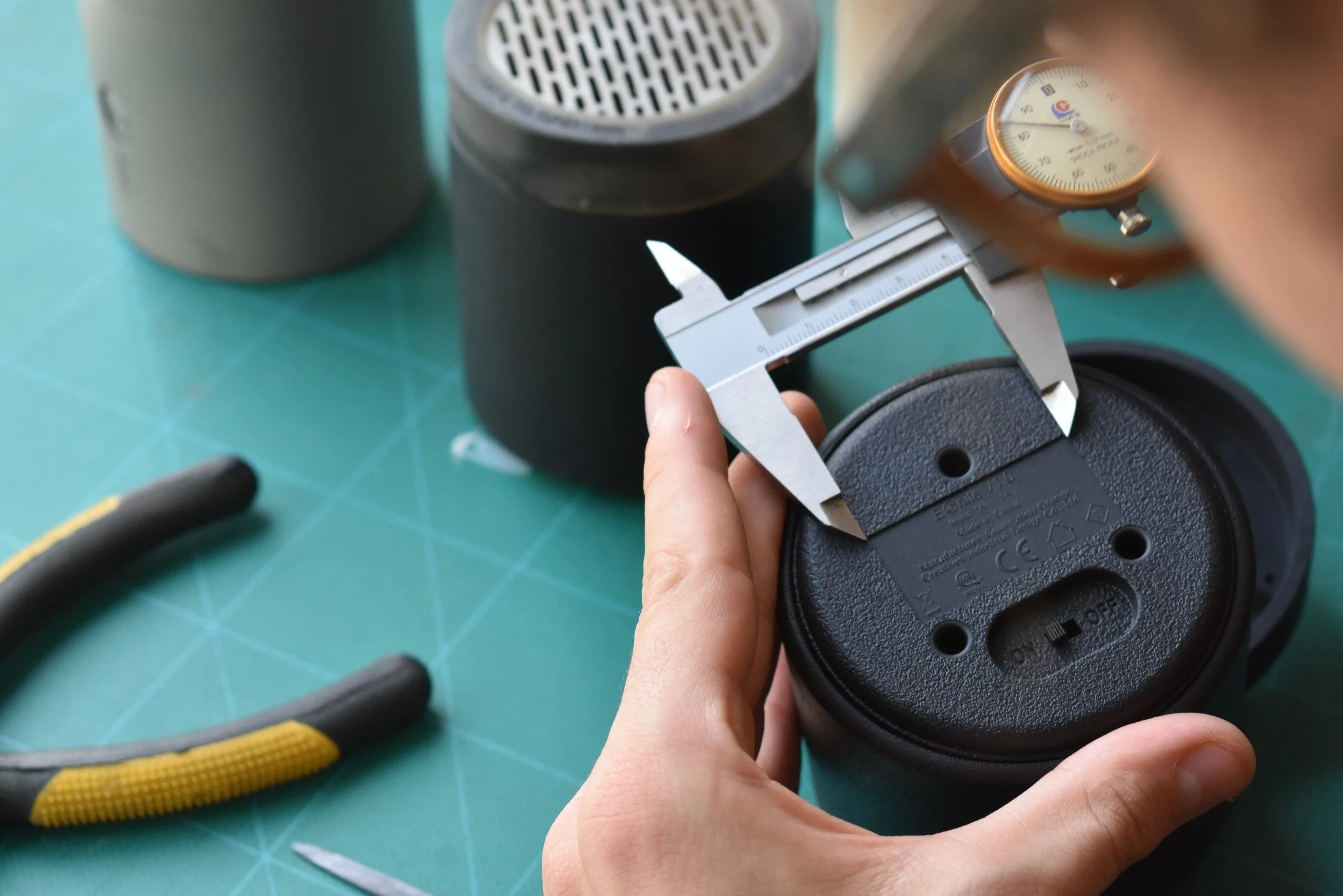 Person measuring the diameter of a black wireless speaker with a digital caliper on a green cutting mat, with tools and a gray cylindrical object nearby.