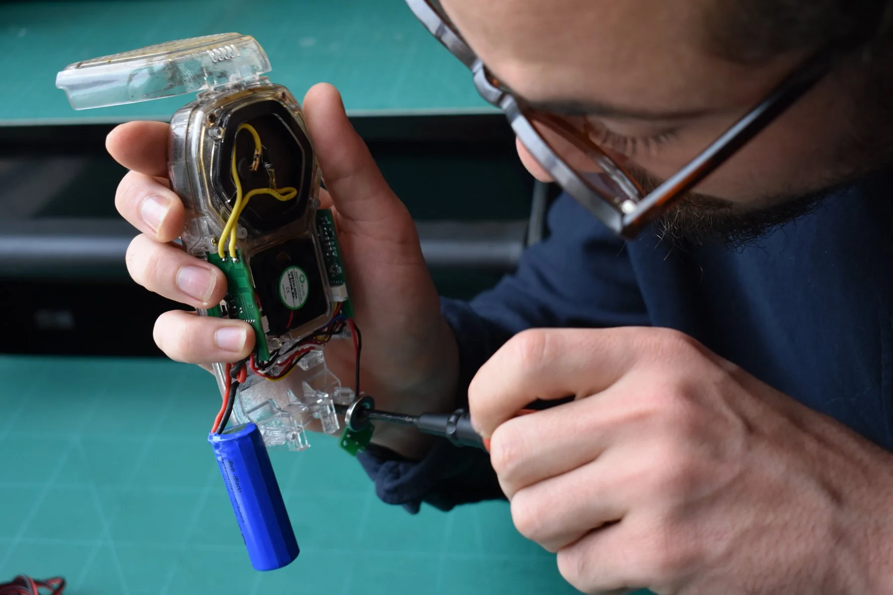 Person wearing glasses working on a small electronic device with circuit boards and wires on a green workbench.