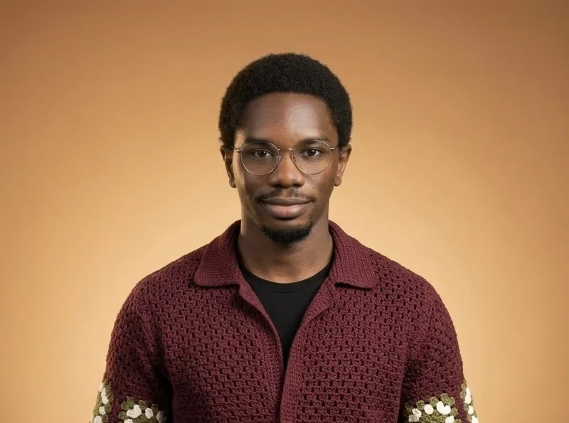 A young man with dark skin, short curly hair, and a trimmed beard and mustache, taking a selfie indoors with a neutral expression. He is wearing a light blue shirt and a thin chain necklace, with a background showing a window with curtains.