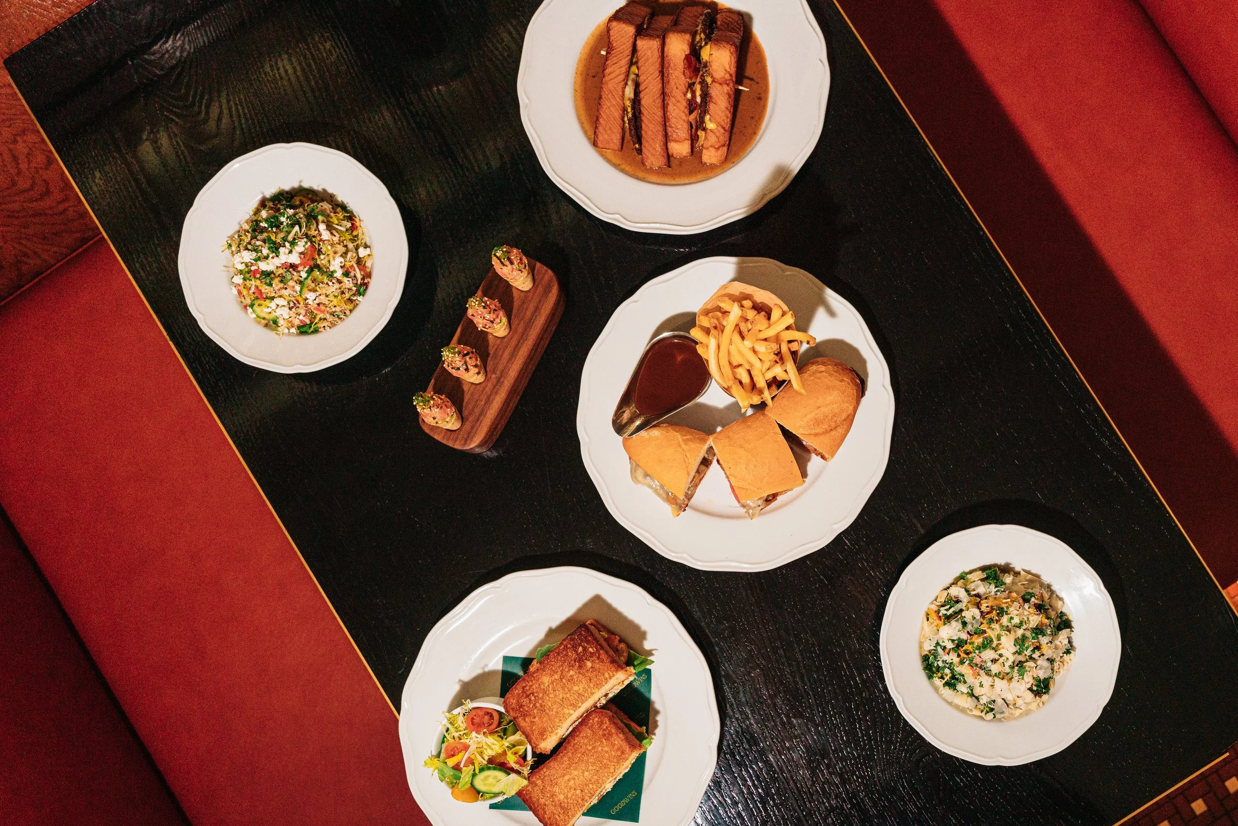 A variety of dishes on a black wooden table, including sandwiches with fries, a salad, a wrap with a side salad, a plate of sushi rolls, a platter of sliders with sauce, and a plate of grilled sandwiches.