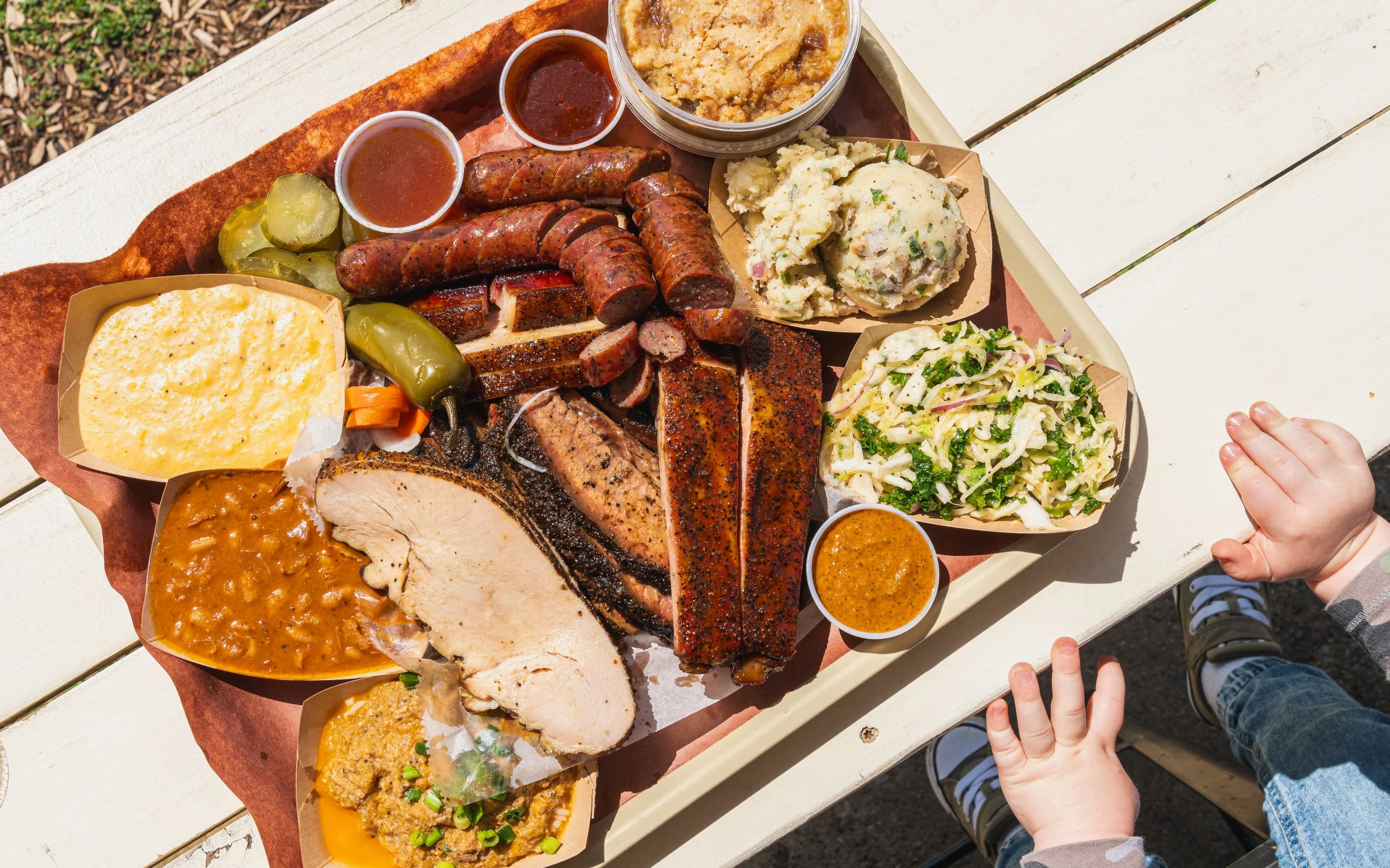 A tray of various barbecue dishes, including sliced brisket, sausages, baked beans, potato salad, coleslaw, mashed potatoes, and green pickles, served on a white table.