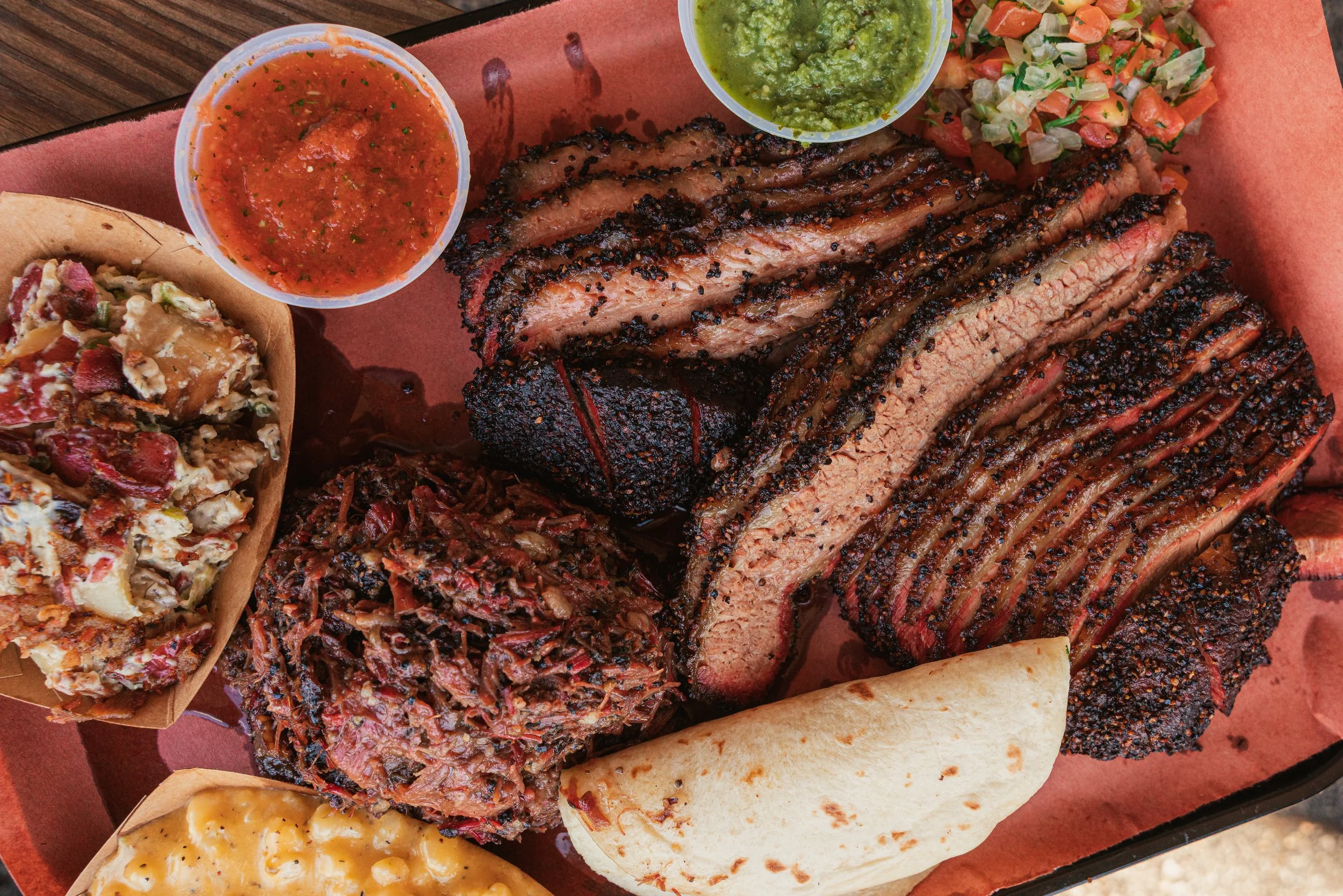 Plate with sliced smoked brisket, barbecue sauce, chopped salsa, cornbread, and side dishes including potato salad, macaroni and cheese, and baked beans.