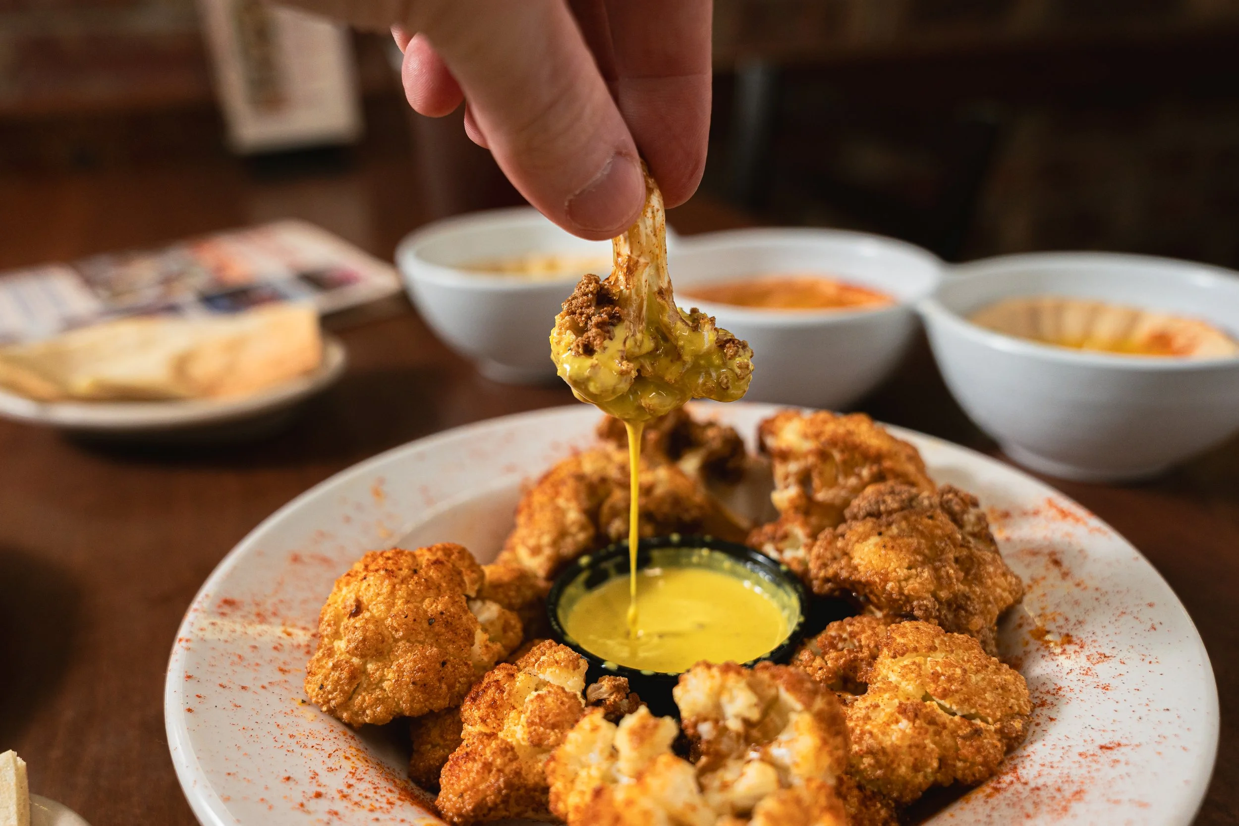 Fried chicken pieces on a plate with a small container of yellow dipping sauce, with a hand pulling a piece of chicken with melted cheese and sauce dripping from it, and three bowls of different sauces in the background.