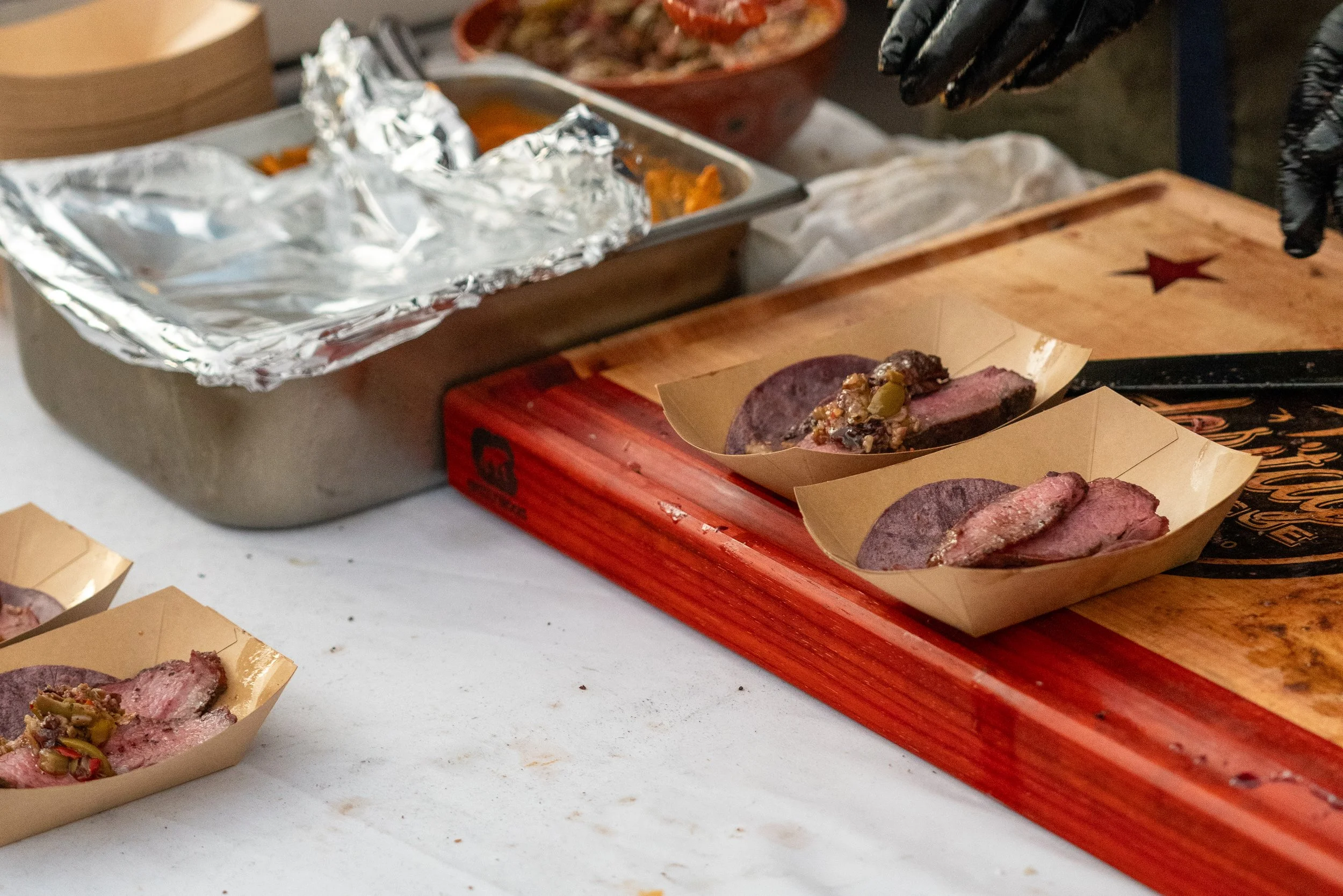 Slices of cooked roast beef with toppings in small paper food cups, on a cutting board with a metal tray and aluminum foil in the background.