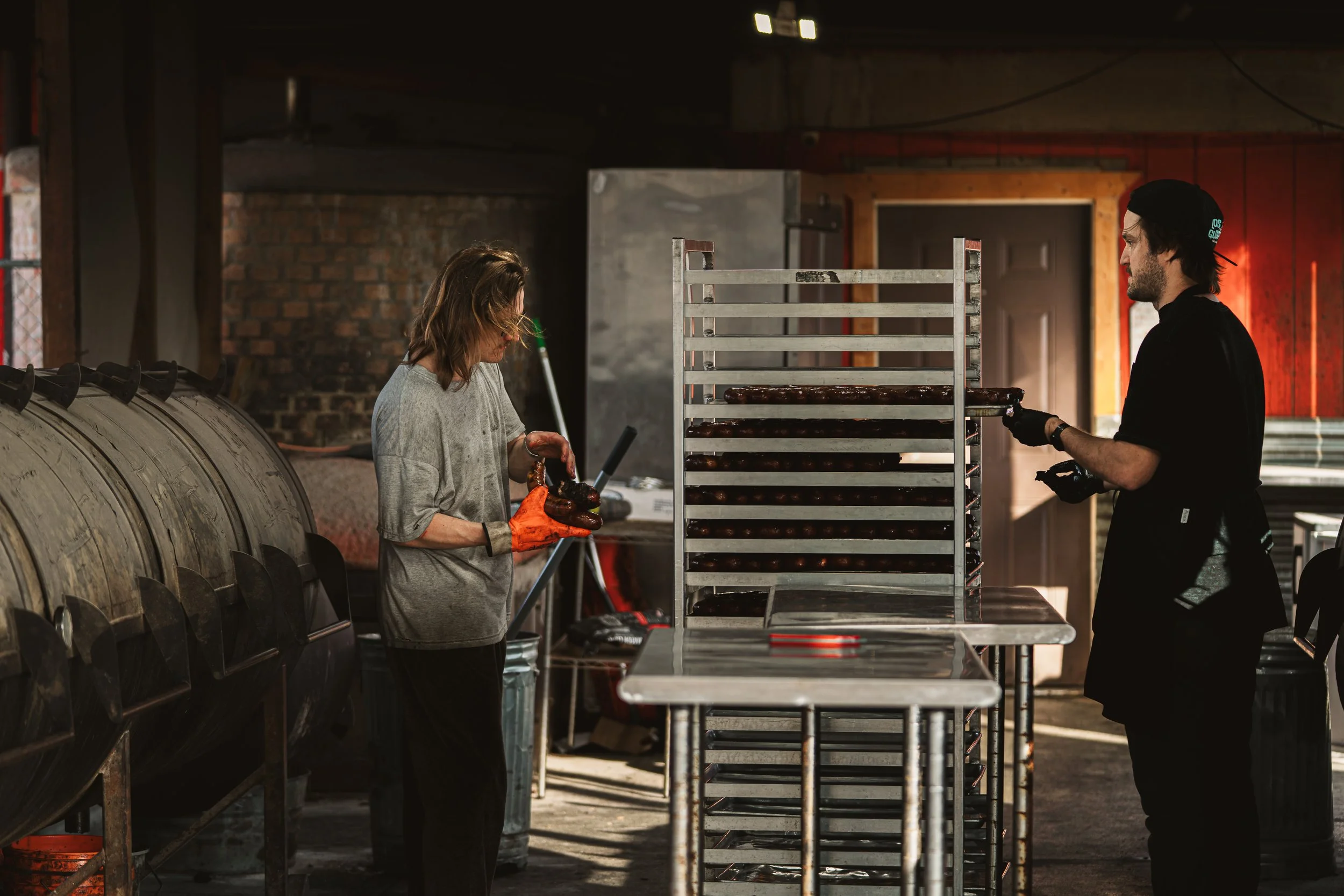 Two people working in a commercial kitchen or bakery, with metal racks and large industrial equipment, preparing or processing food, wearing gloves.