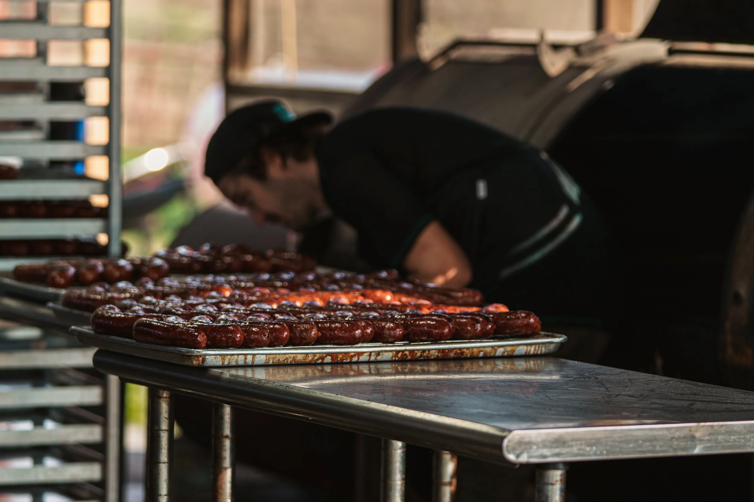 A person leaning over a table of cooking sausages in front of a large smoker or grill, with a rack of more sausages nearby.
