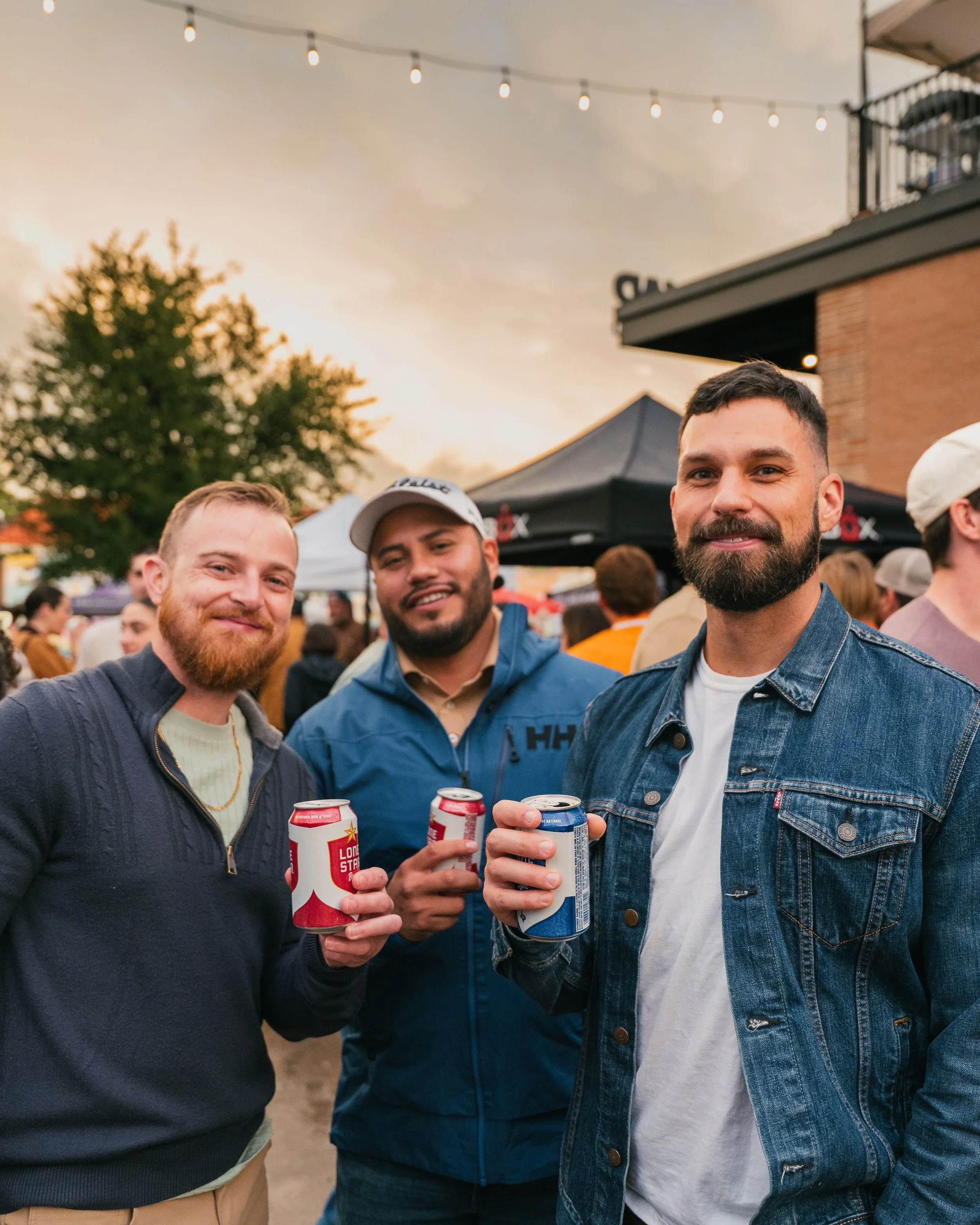 Three men smiling and holding cans of beer at an outdoor event during sunset, with a crowd and tents in the background.