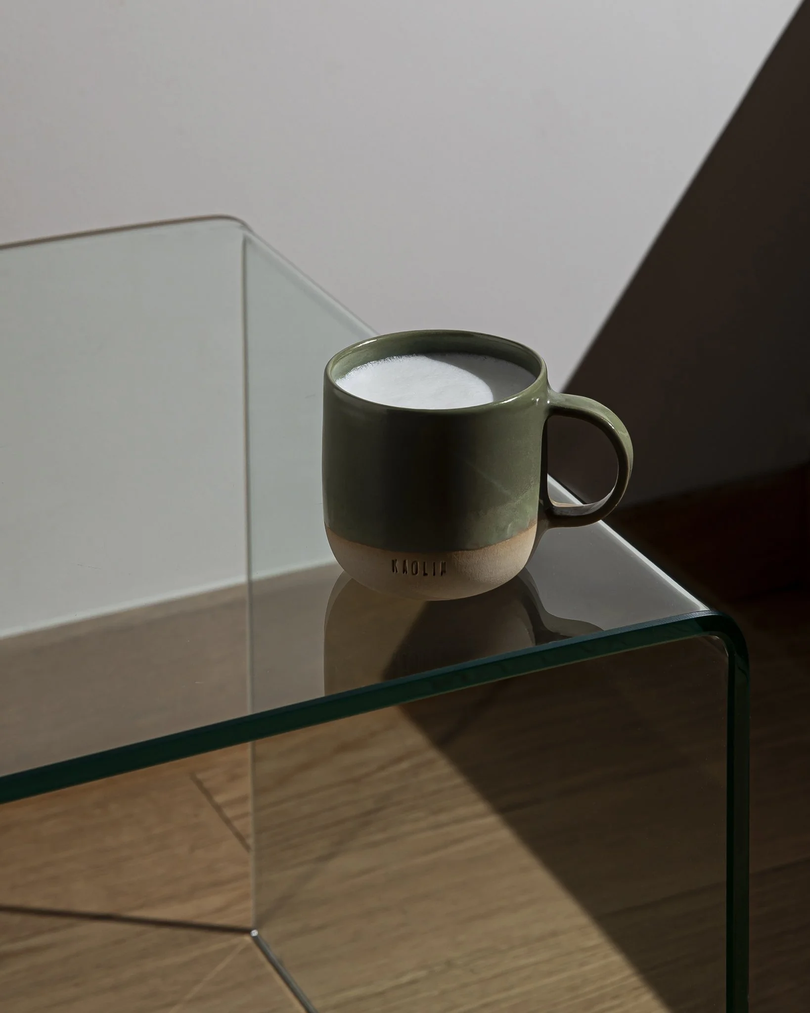 A glossy olive-green Kaolin Ceramics mug filled with milk foam sits on the corner of a clear glass side table, lit by sharp natural sunlight that creates clean geometric shadows across the glass, wall, and wooden floor.