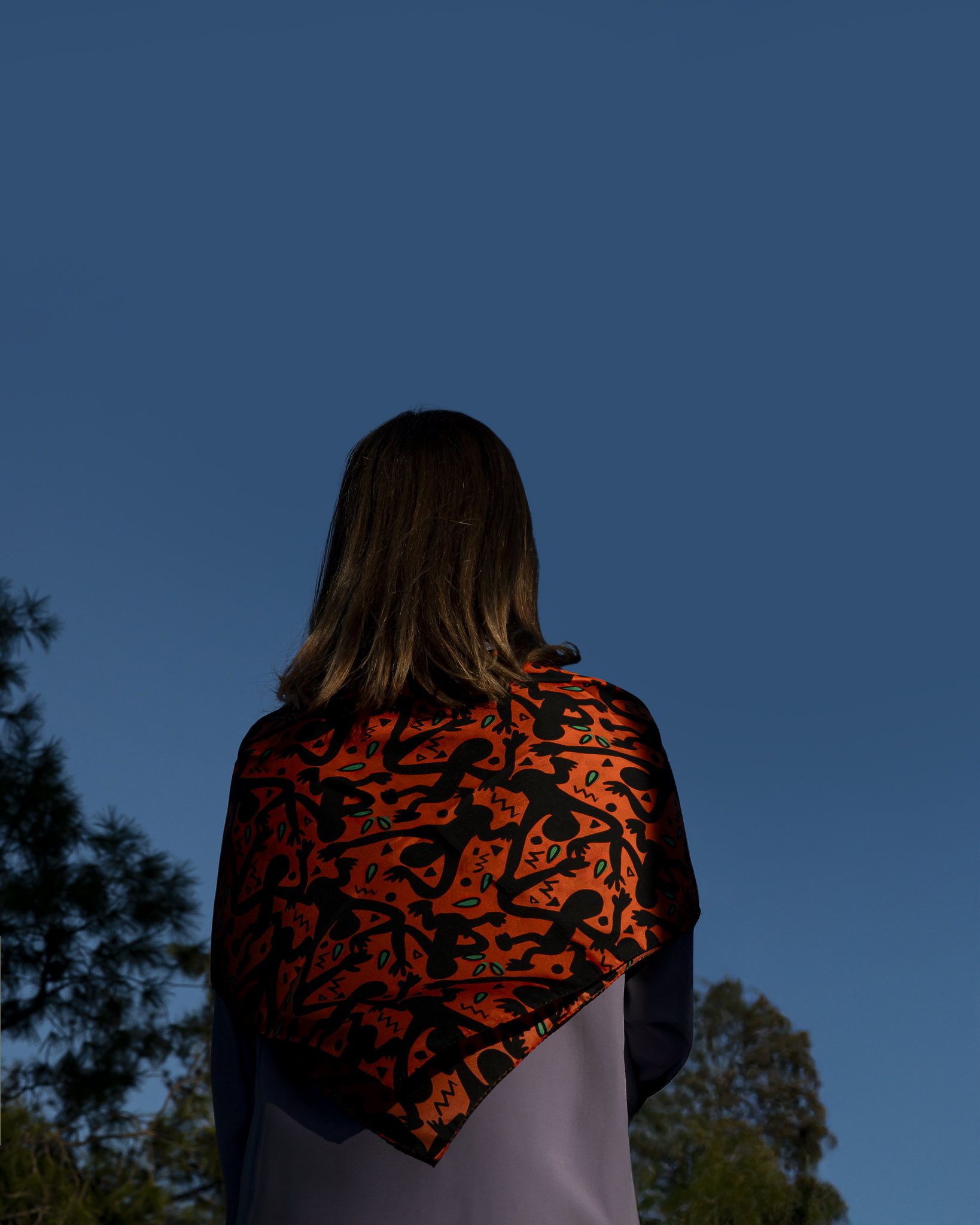 A woman is photographed from behind under a deep blue sky, wearing a bold orange patterned scarf draped over her shoulders, with dark graphic motifs and small green accents, set against a quiet outdoor landscape.
