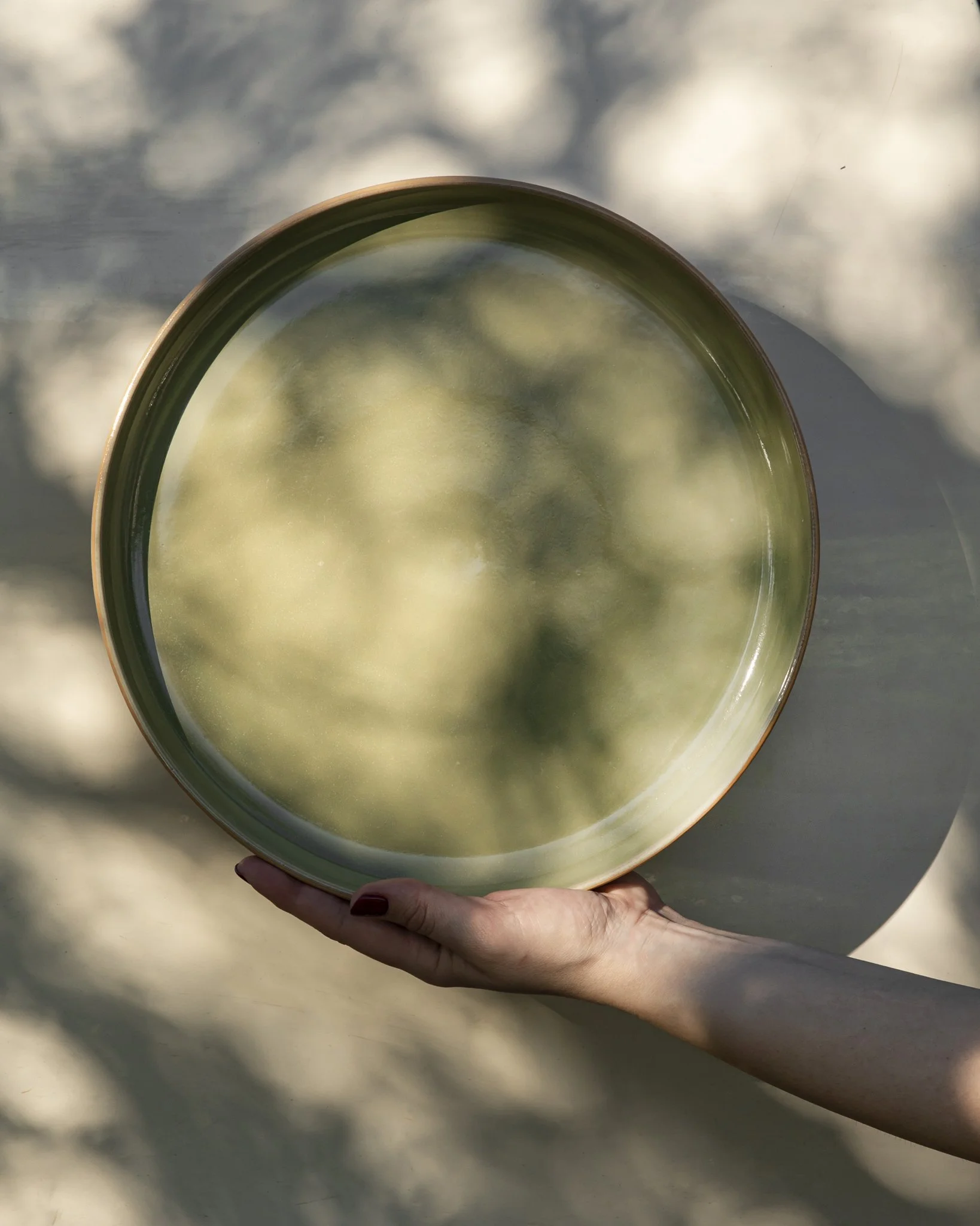 A hand holds a wide, glossy Kaolin Ceramics plate in deep olive glaze, photographed in dappled sunlight that reflects soft, organic shadows across the surface and the neutral background.