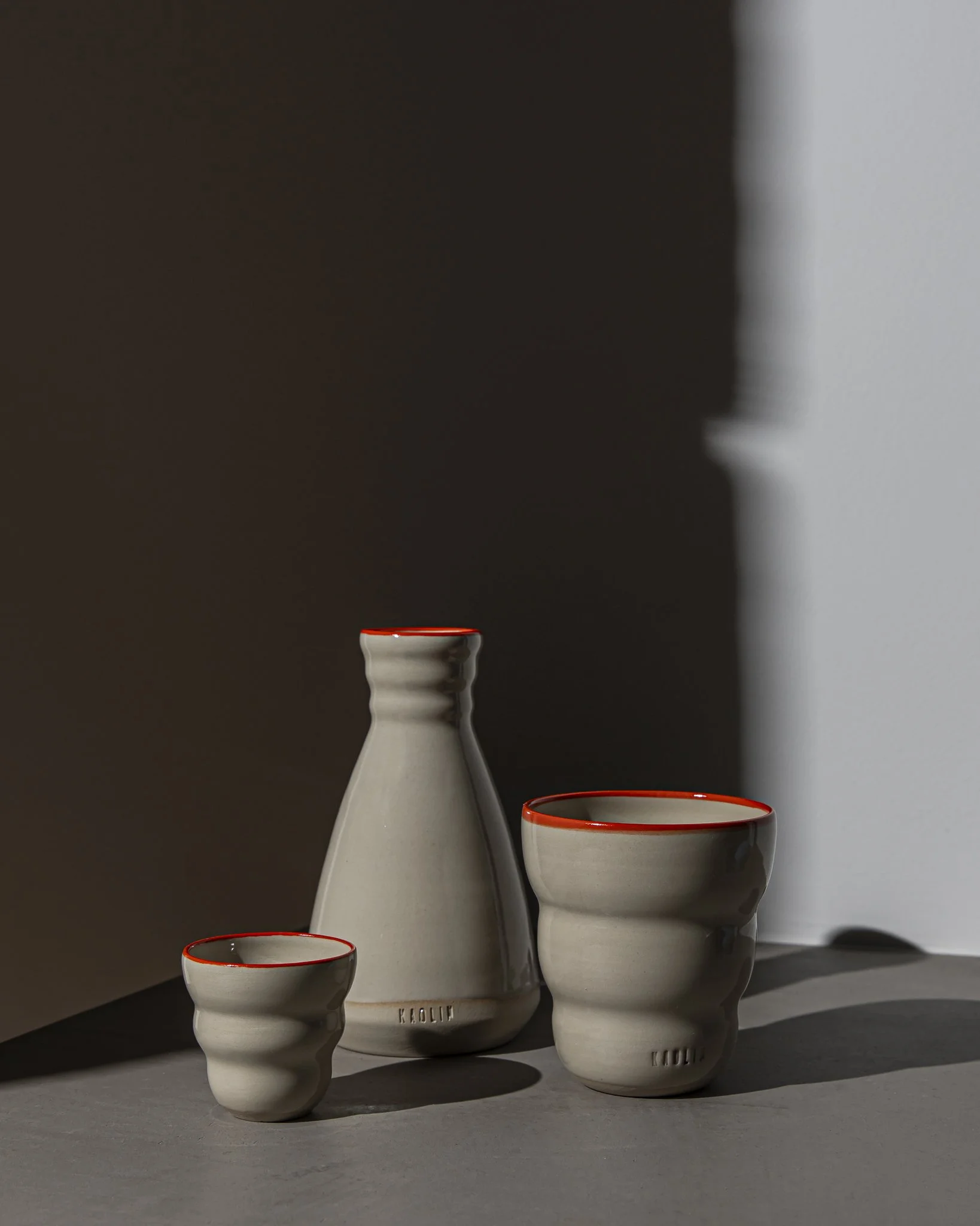 Photography of a set of three white ceramic bowls with orange rims on a gray surface, casting shadows, against a dark and light background.