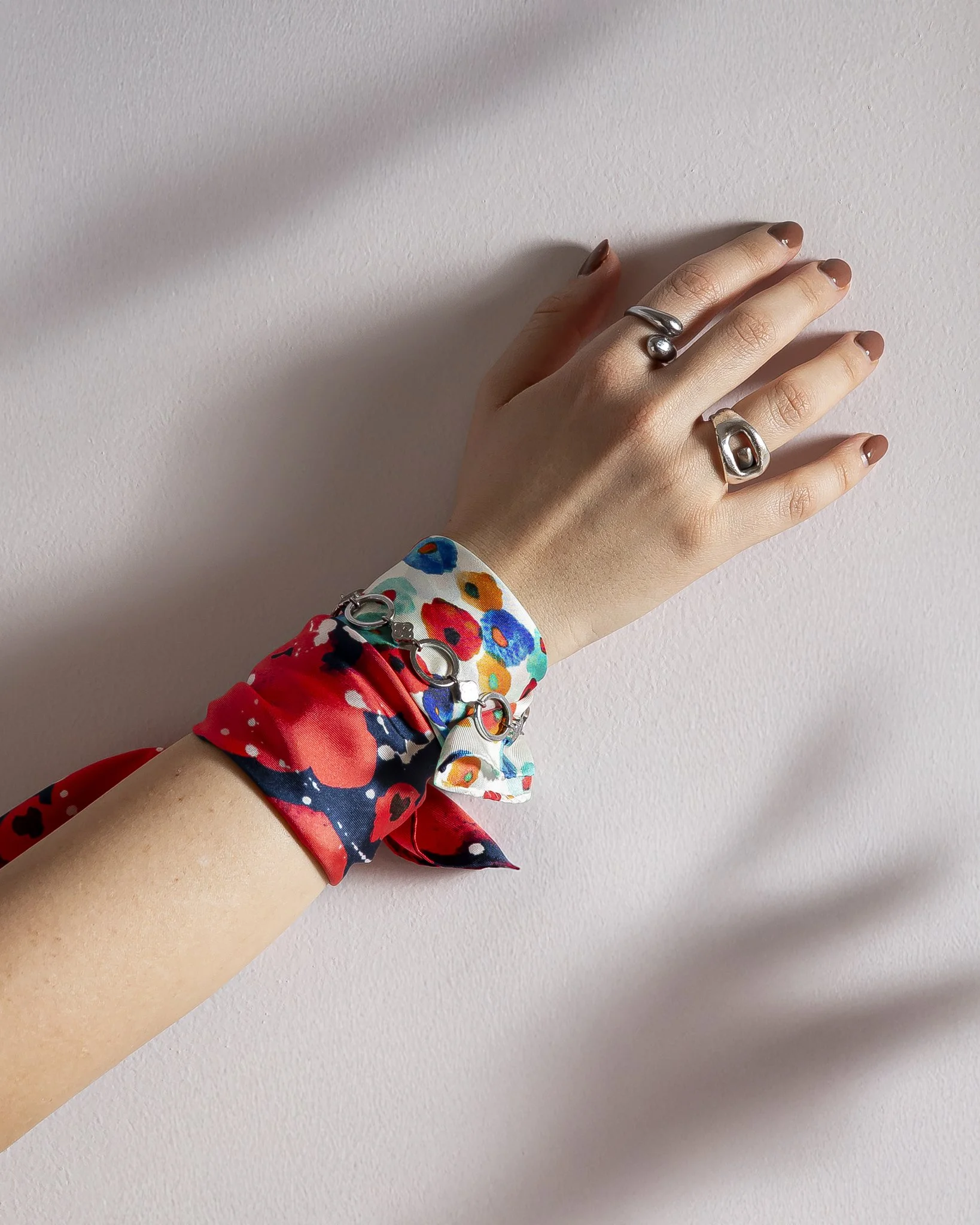 A close-up of a woman’s hand with silver rings and neutral nails, wearing a vibrant silk scarf bracelet in red, navy, and multicolor floral print with small chain details, photographed in soft natural light against a pale textured wall.