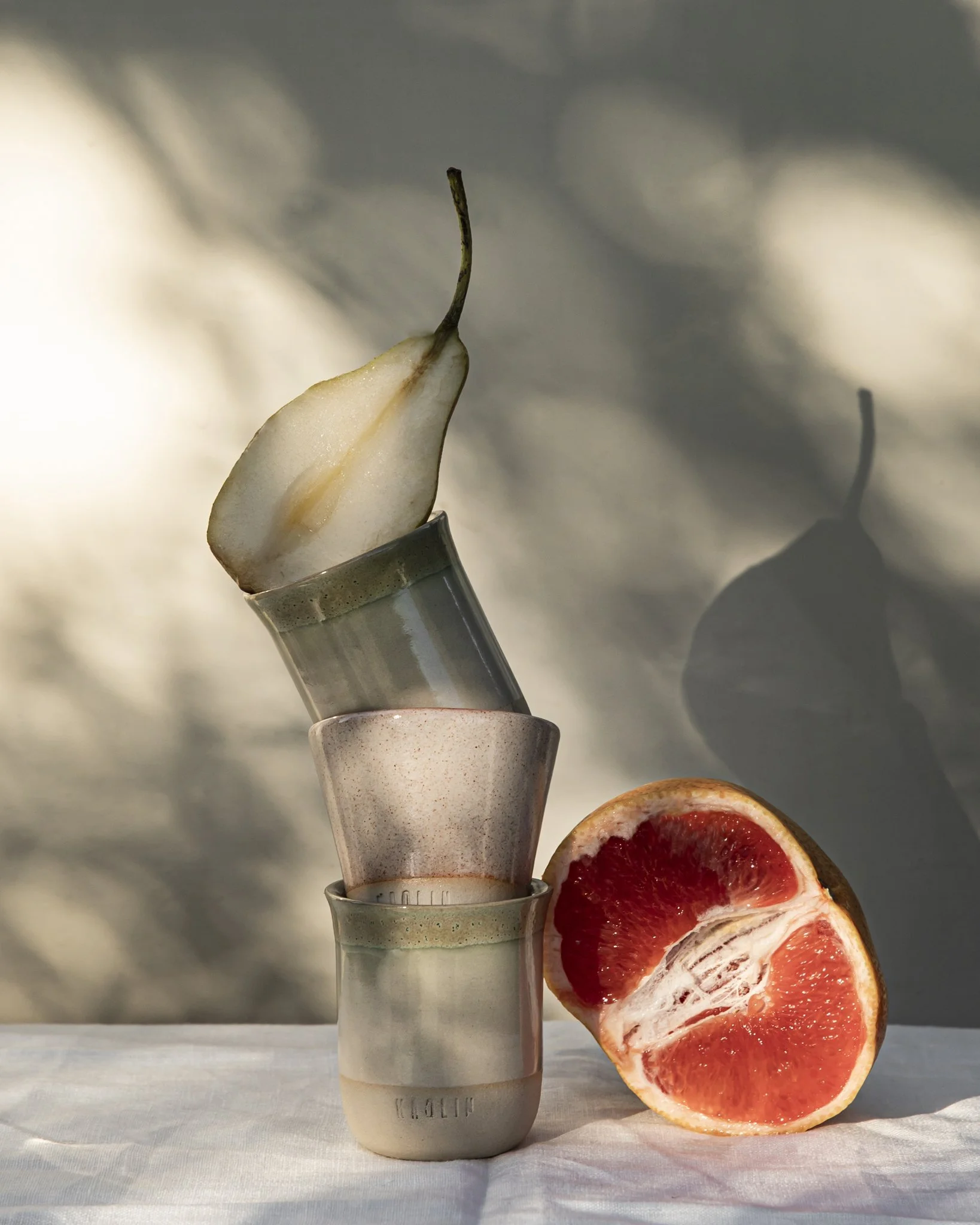 A sculptural still life featuring three stacked Kaolin Ceramics cups in muted green and speckled stone glazes, crowned with a pear slice and paired with a halved grapefruit, photographed on linen in dappled natural light with soft, botanical shadows.