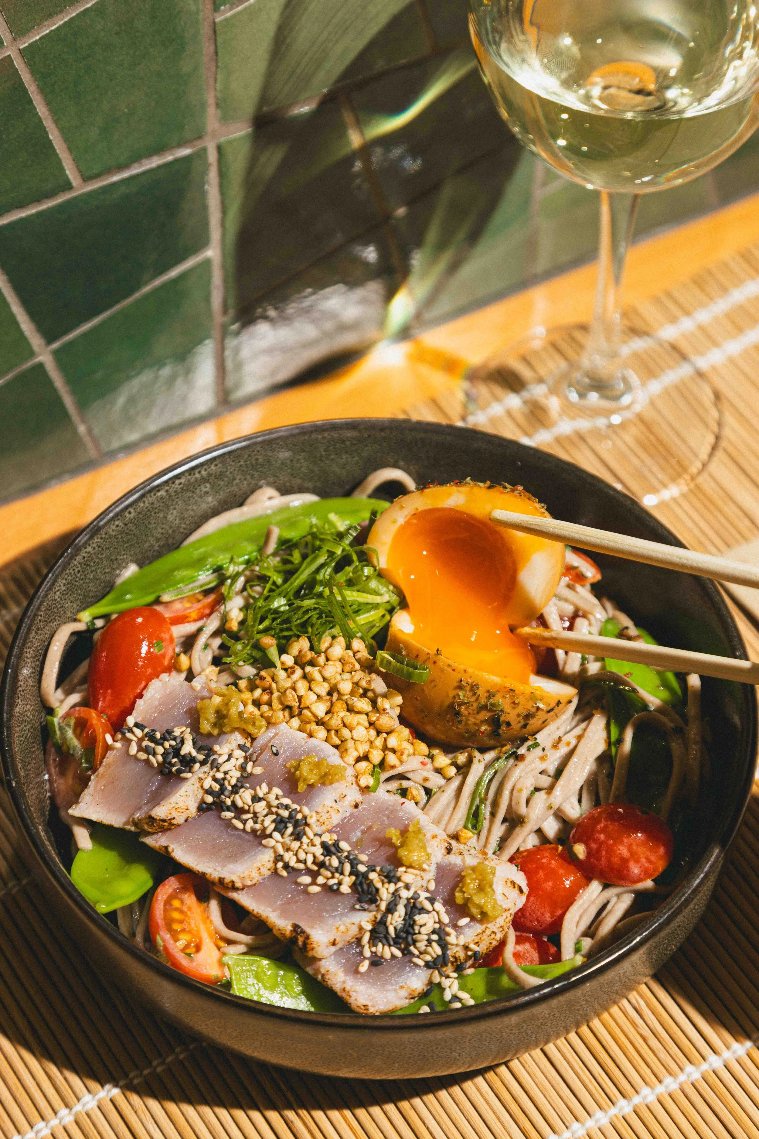 A bowl of Asian-style noodles topped with sliced meat, cherry tomatoes, snow peas, a soft-boiled egg, green onions, chopped nuts, and sesame seeds, with a glass of white wine nearby.