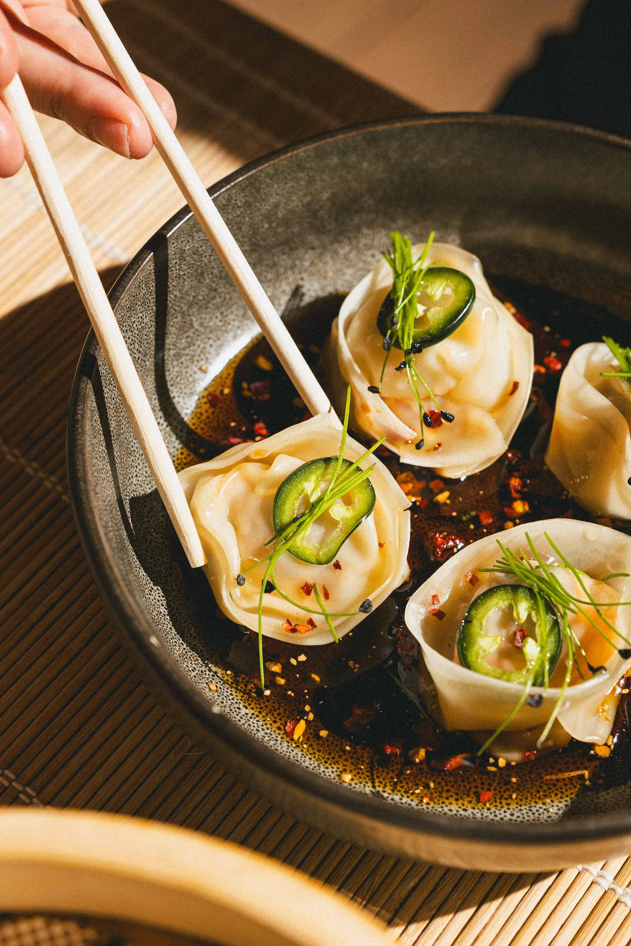 Close-up of steamed dumplings in a dark bowl, garnished with sliced jalapenos and microgreens, served in a soy-based dipping sauce with chili flakes.