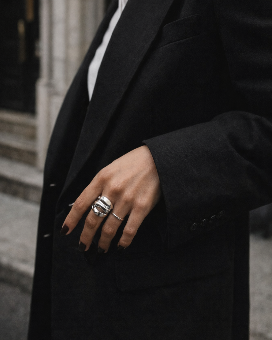 Close-up of a person wearing a black blazer, with their hand visible adorned with silver rings and dark nail polish, in an urban setting.