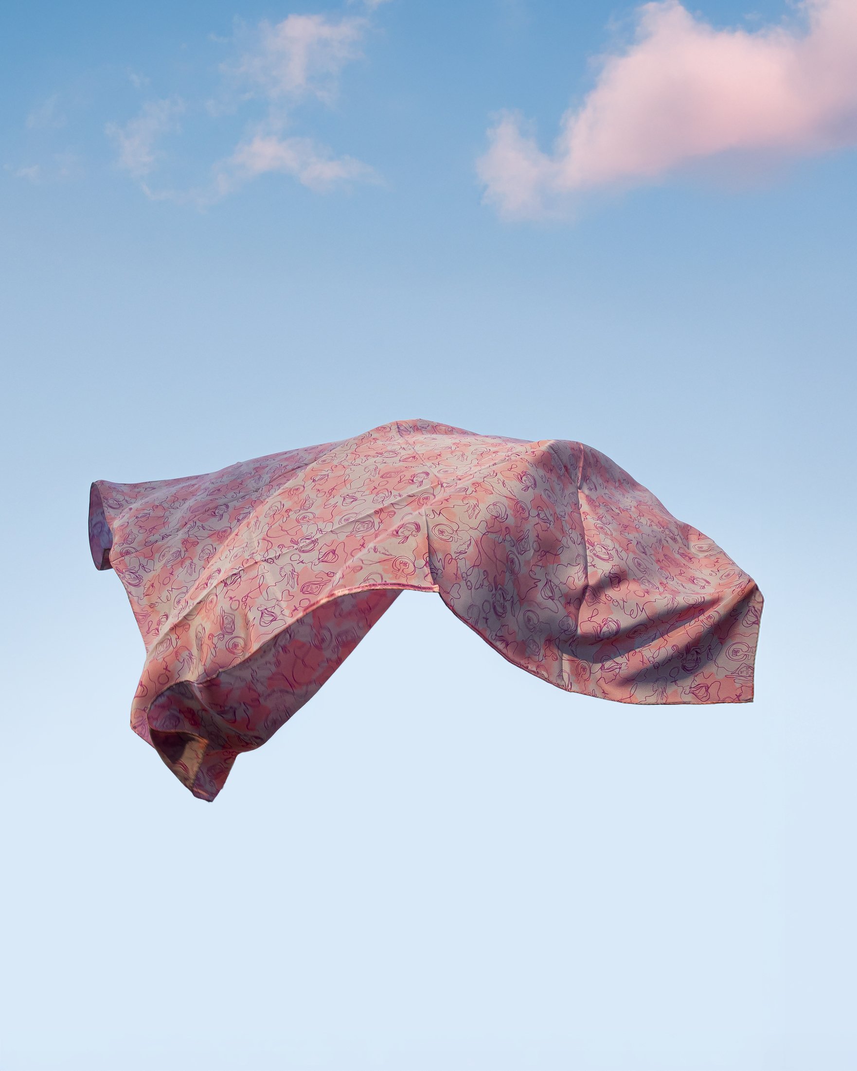 A pink patterned silk scarf floats midair against a clear blue sky with soft clouds, captured in motion as the fabric billows and catches the light.