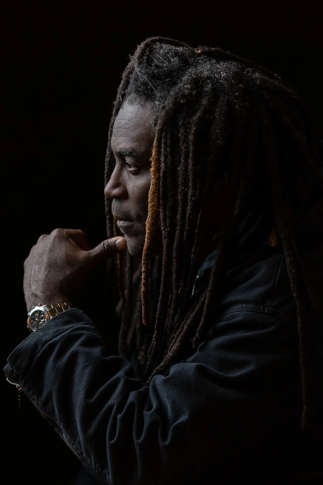 A side profile of a man with long dreadlocks wearing a black jacket and a watch, posing against a dark background.