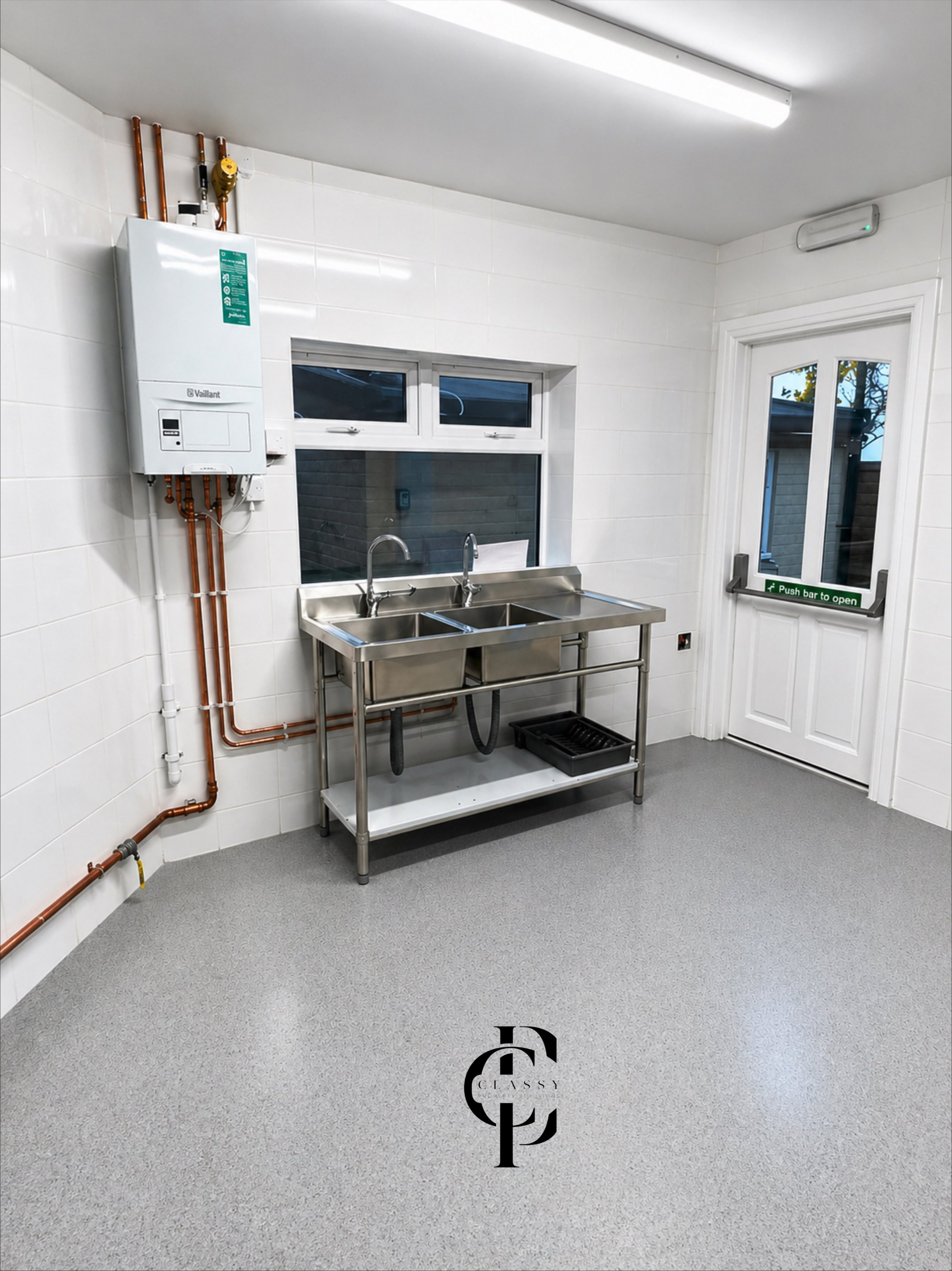 A clean, white kitchen or utility room with a stainless steel sink and drainboard, a window above it, a white door with a push bar to open, and an electric water heater on the wall. There is a black tray or mat on the floor beneath the sink.