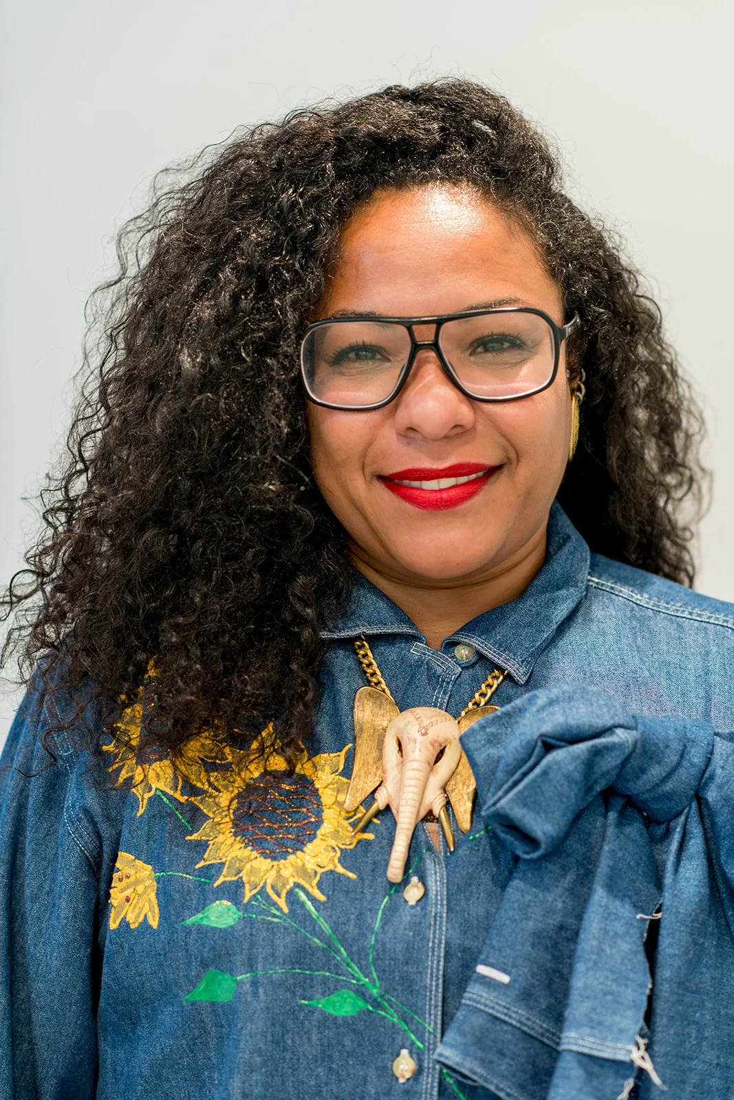 A woman with shoulder-length brown hair, wearing a dark blue denim shirt, a gold necklace, and glasses, standing and smiling at the camera against a plain background.