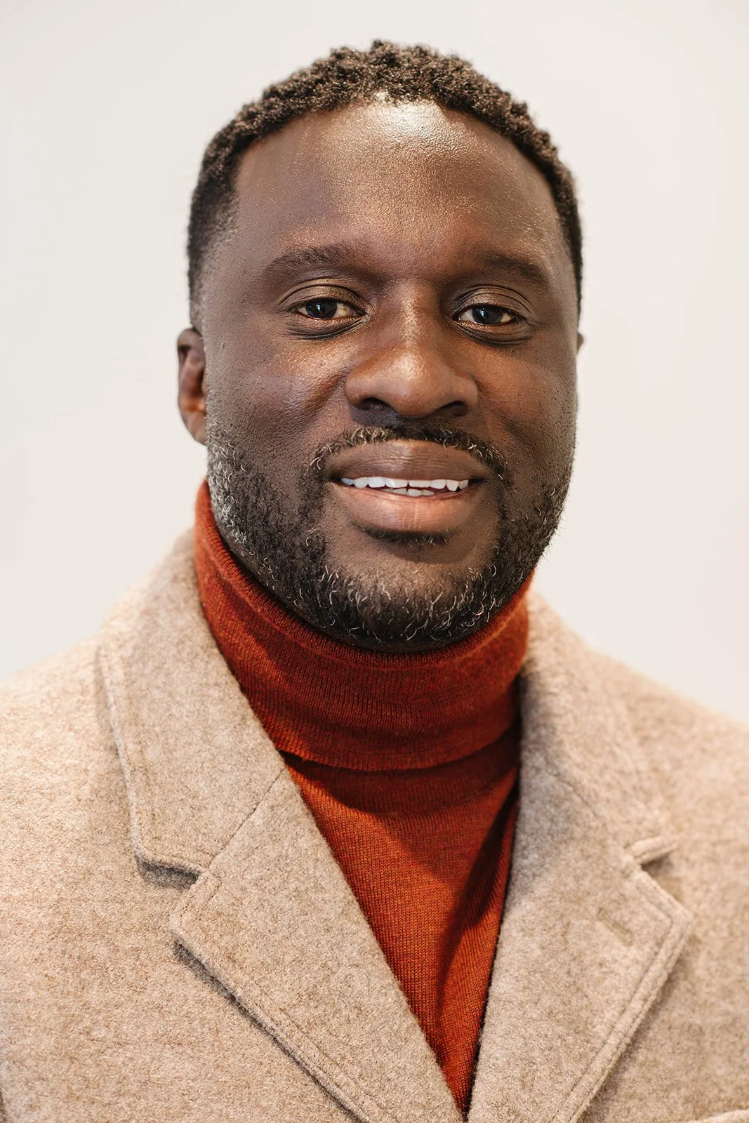 Portrait of a Black man wearing an orange turtleneck and a cream jacket, against a plain neutral background.