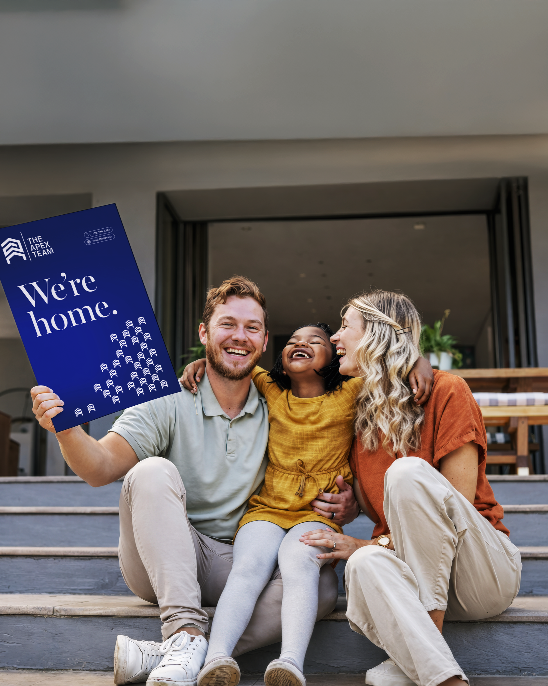 Happy clients hold a branded Apex Team "We're home." sign outside their new front door, celebrating a successful real estate purchase.