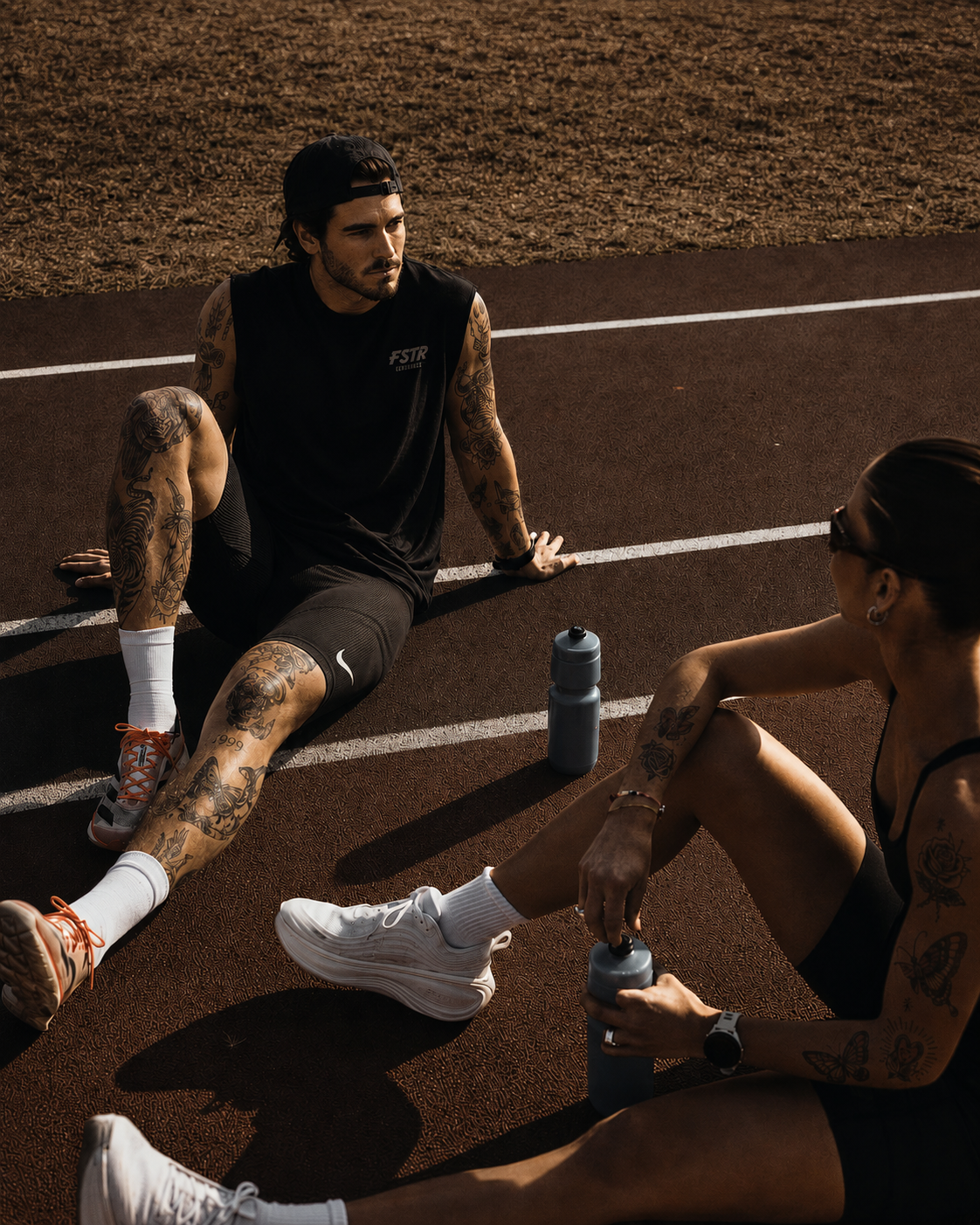 FSTR Endurance brand identity in use — two athletes resting on a running track wearing FSTR Endurance apparel, demonstrating the logo applied to athletic wear.