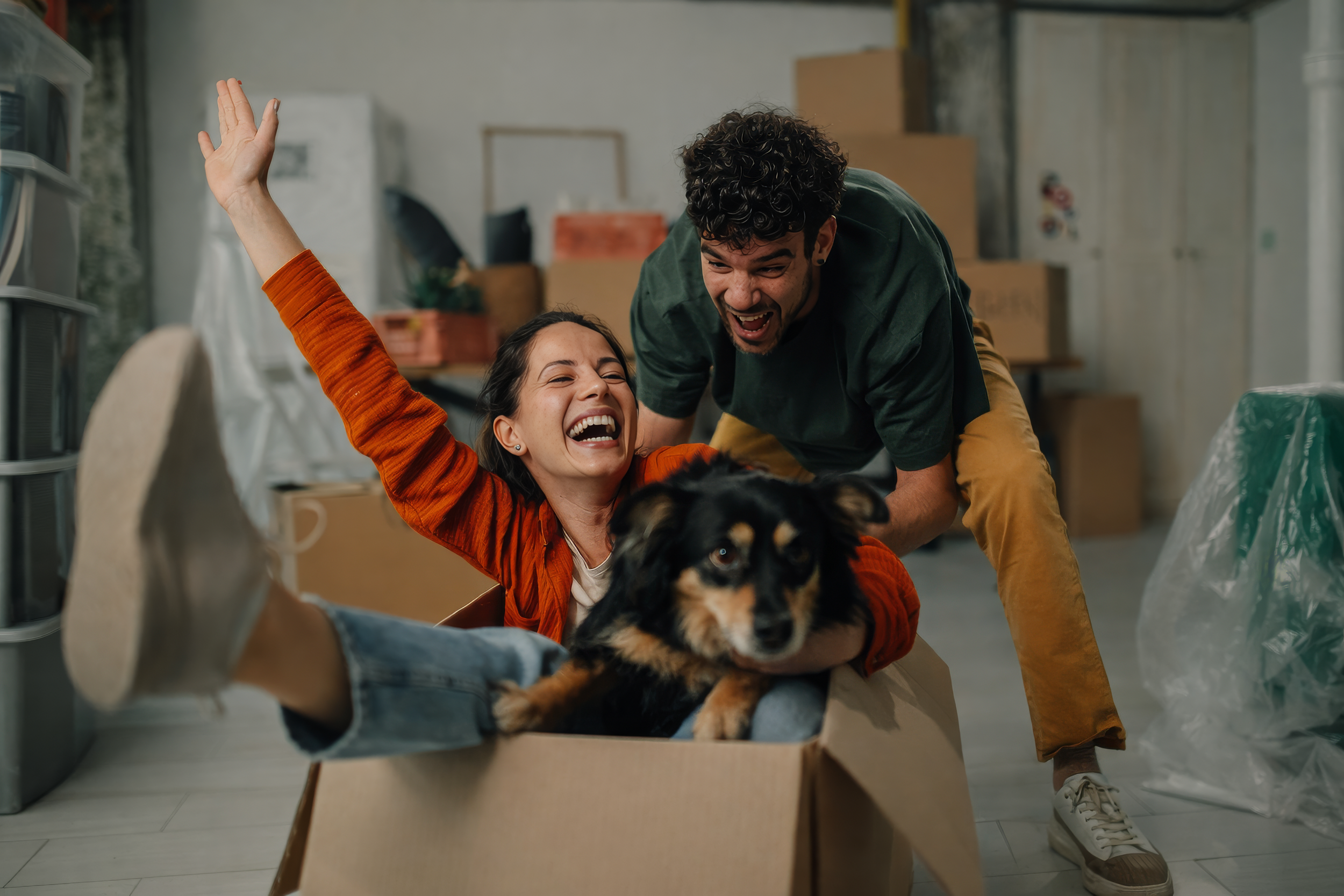 Laughing couple celebrate moving day — woman sits in a cardboard box with a dog while her partner pushes her through their new home.