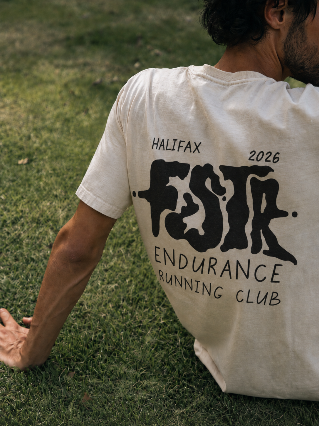 FSTR Endurance limited-edition drip logo on a white T-shirt — specialty brand identity design with hand-done melted letterforms, worn by a running club member seated on grass.