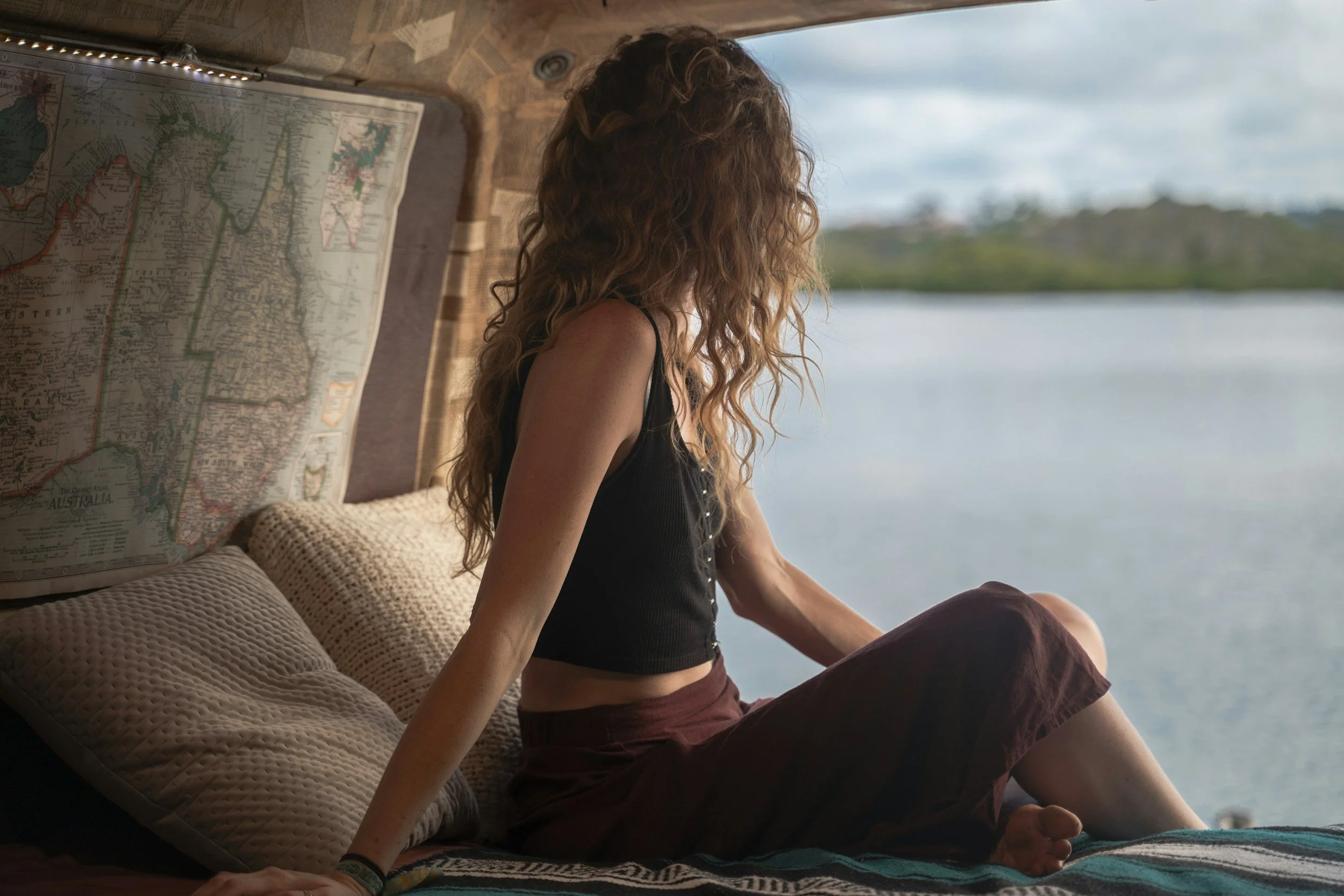 Une femme aux cheveux bouclés roux, assise sur un lit à l'intérieur d'une cabane en bois, regardant par la fenêtre vers un lac ou un fleuve.