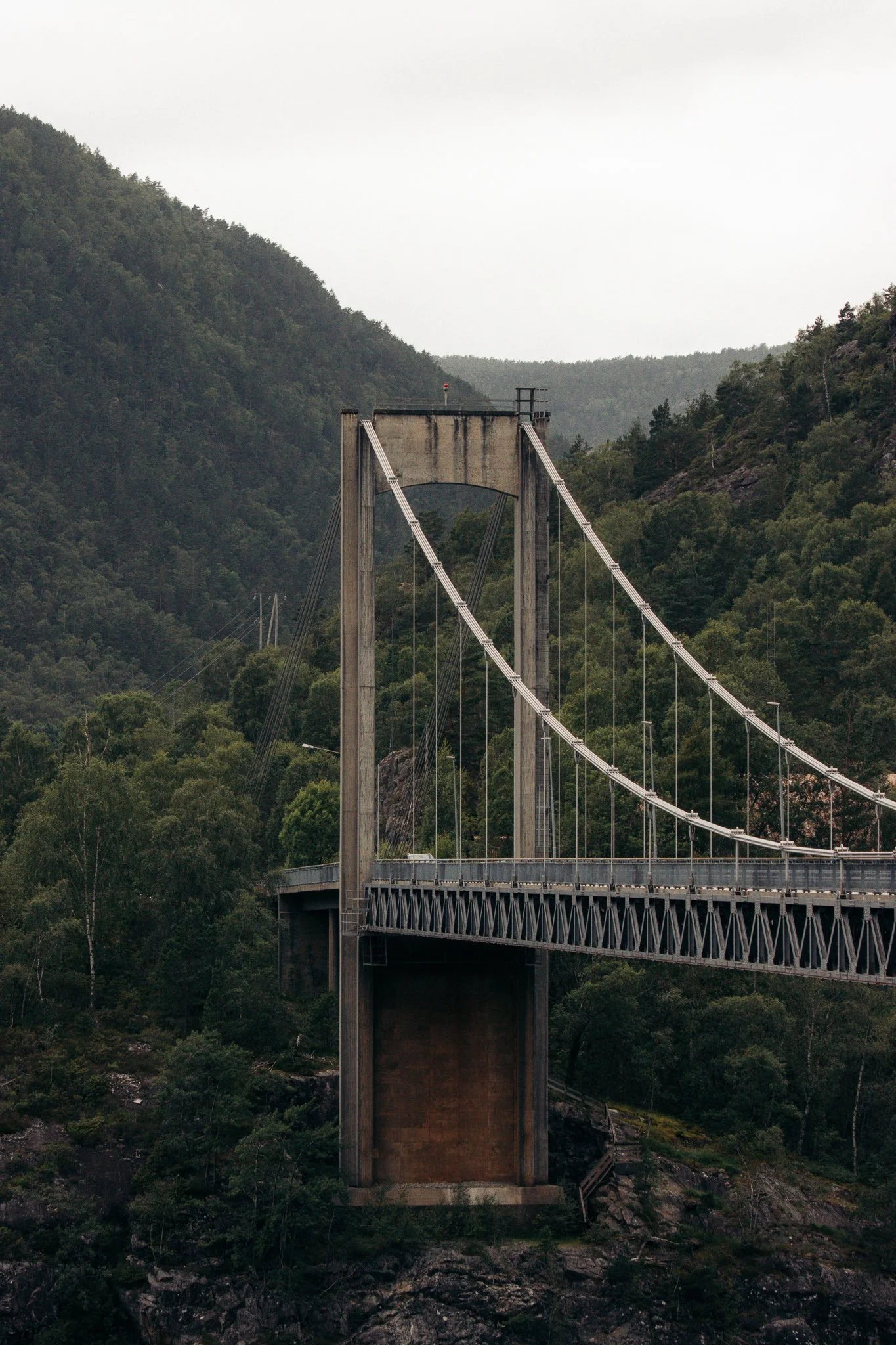 A suspension bridge with tall concrete towers spans across a forested mountainous landscape, under an overcast sky.