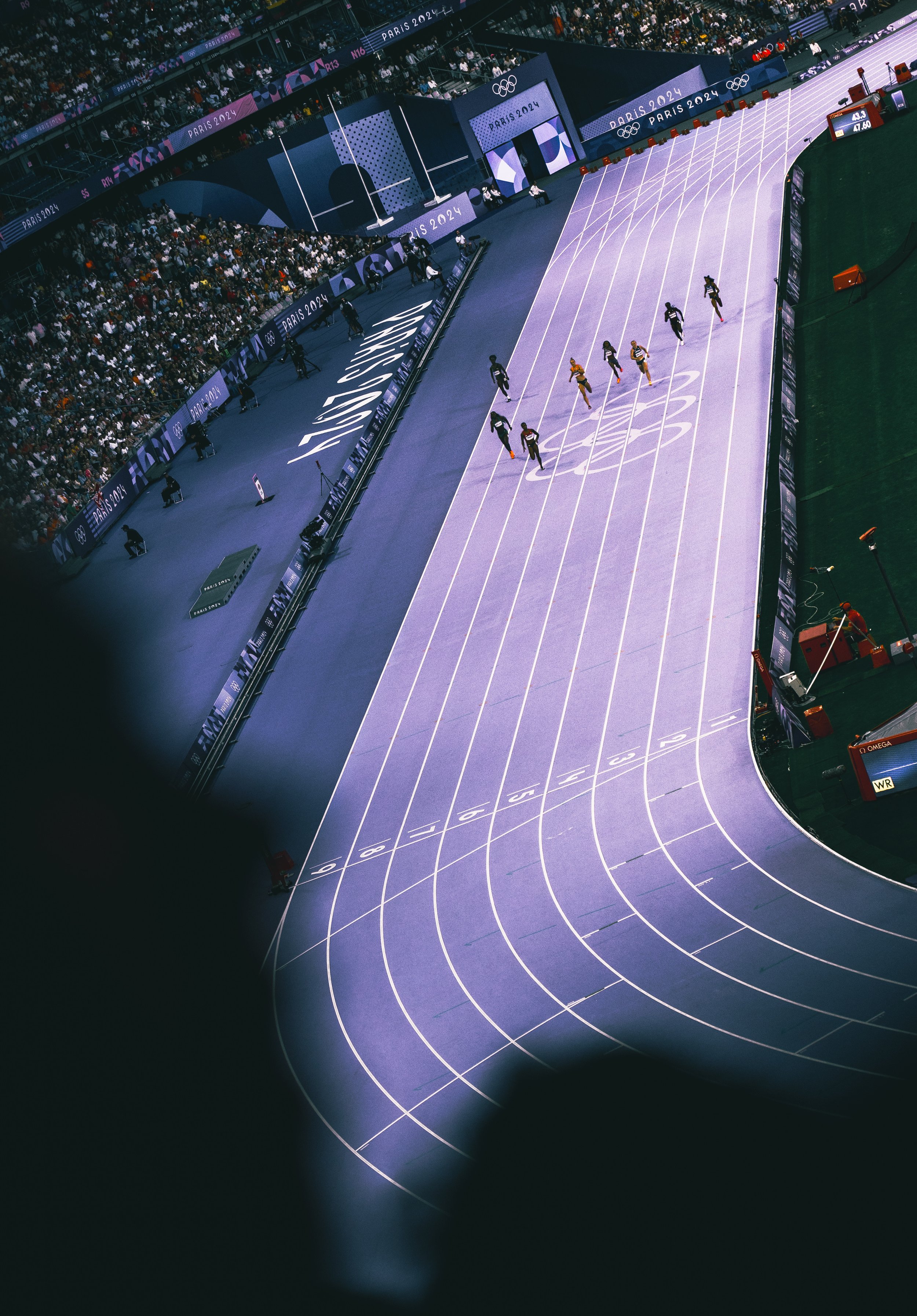Indoor track and field stadium with athletes competing in a race, audience in stands, and Olympic banners.